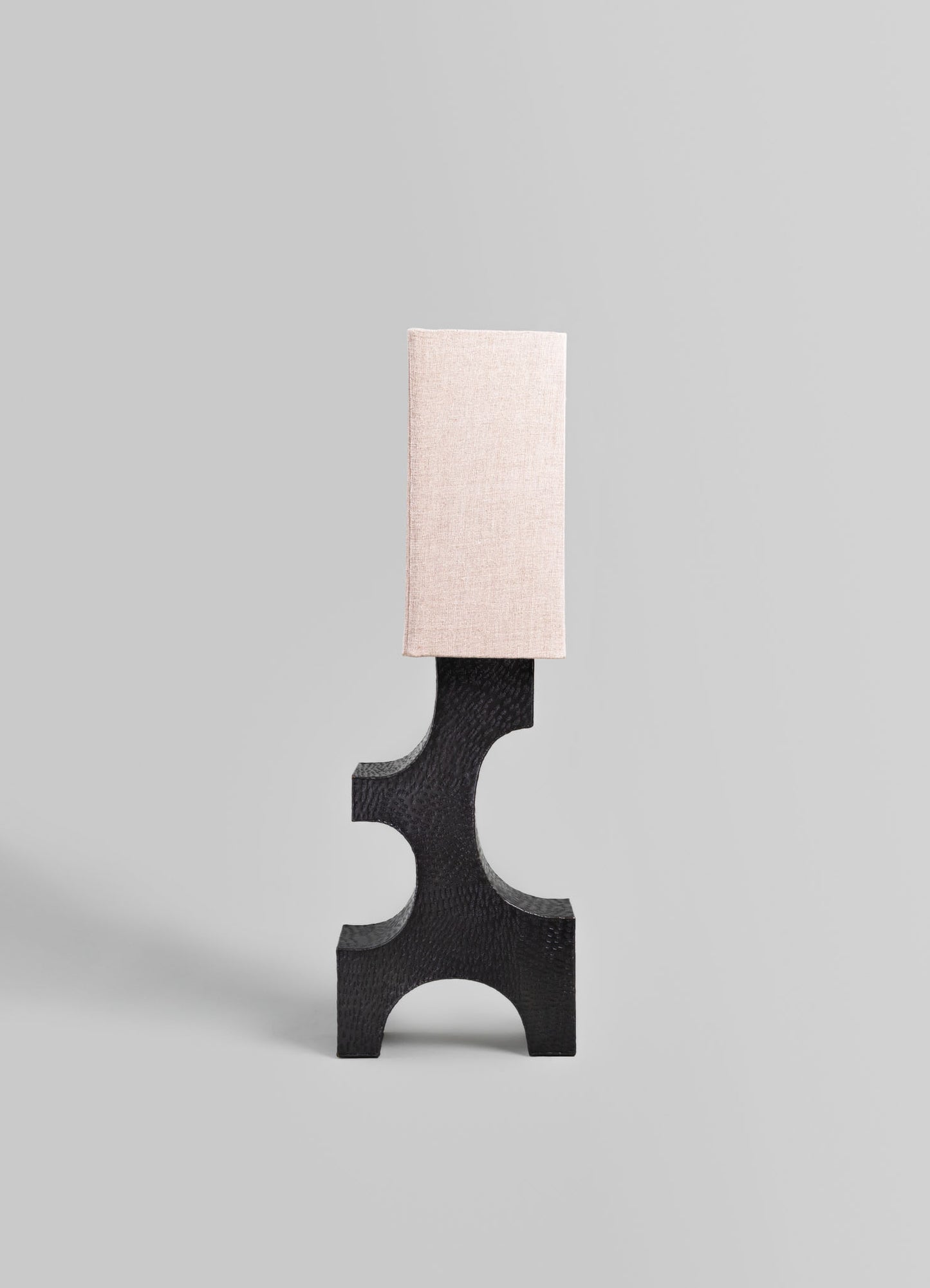 A table lamp with a black textured base featuring geometric cutouts and a rectangular fabric lampshade in a soft beige tone, set against a neutral background.