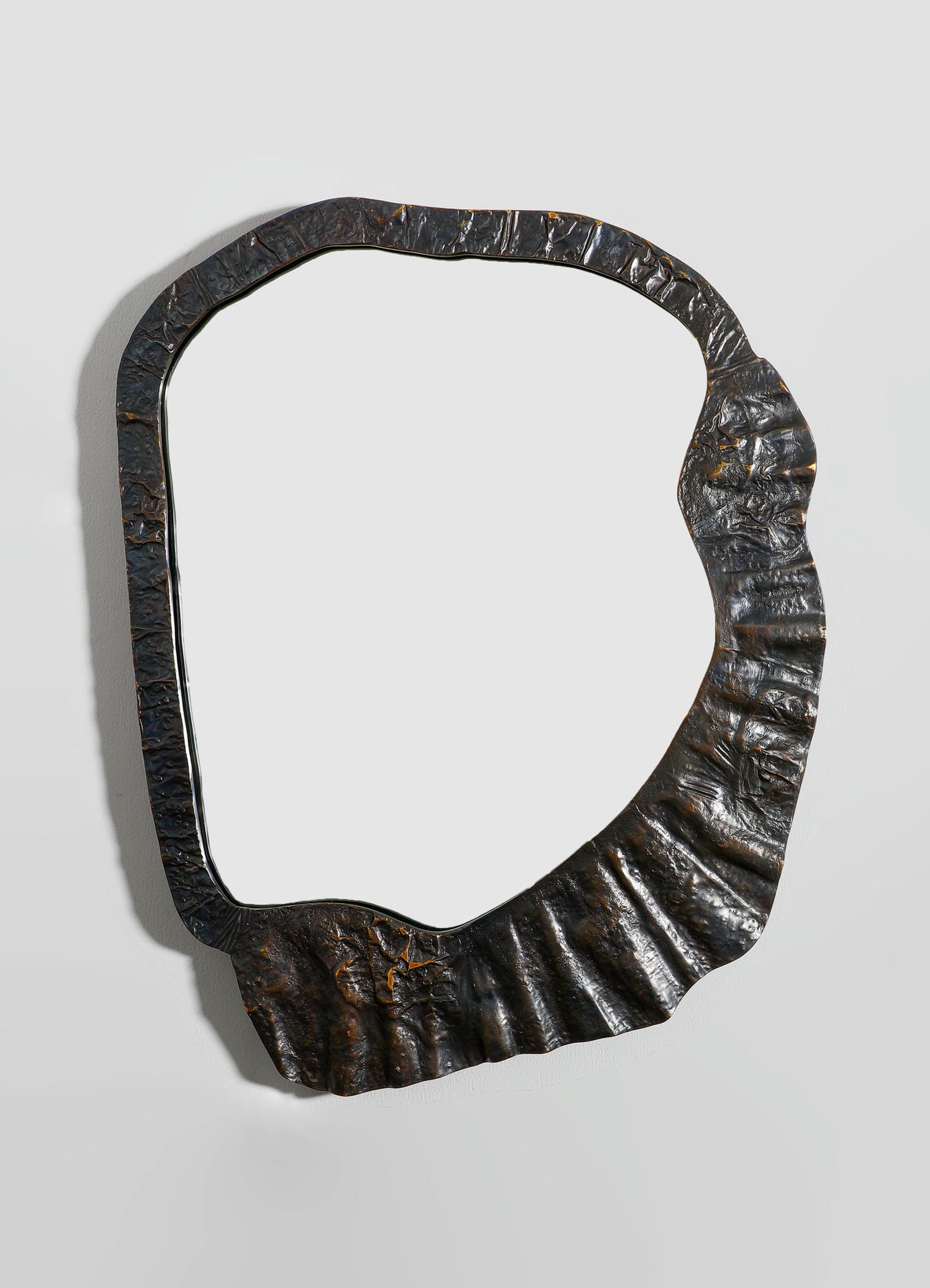 A decorative mirror with a frame made of textured, dark metal with a hammered finish, featuring an irregular, organic shape. The mirror's style is contemporary with a rustic, industrial aesthetic.