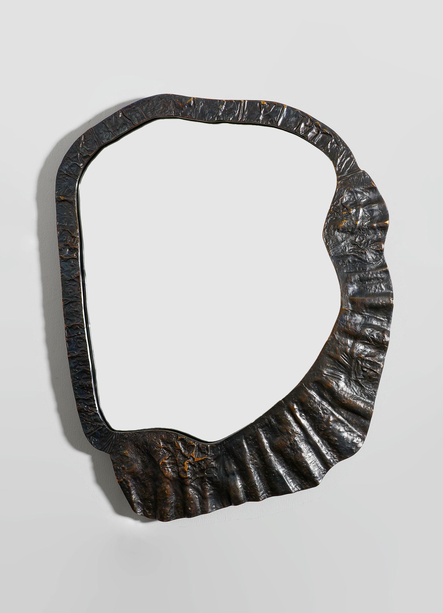 A decorative mirror with a frame made of textured, dark metal with a hammered finish, featuring an irregular, organic shape. The mirror's style is contemporary with a rustic, industrial aesthetic.
