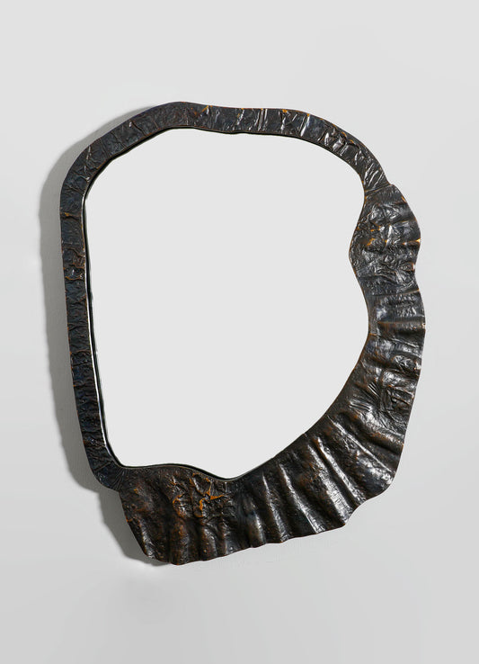 A decorative mirror with a frame made of textured, dark metal with a hammered finish, featuring an irregular, organic shape. The mirror's style is contemporary with a rustic, industrial aesthetic.