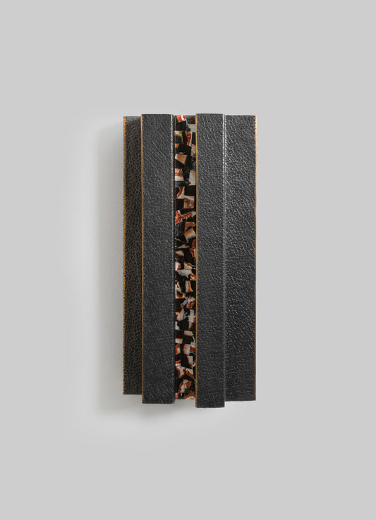 A decorative object featuring black textured leather strips with a central motif of multicolored abstract stone or resin inlay, mounted on a light gray background.