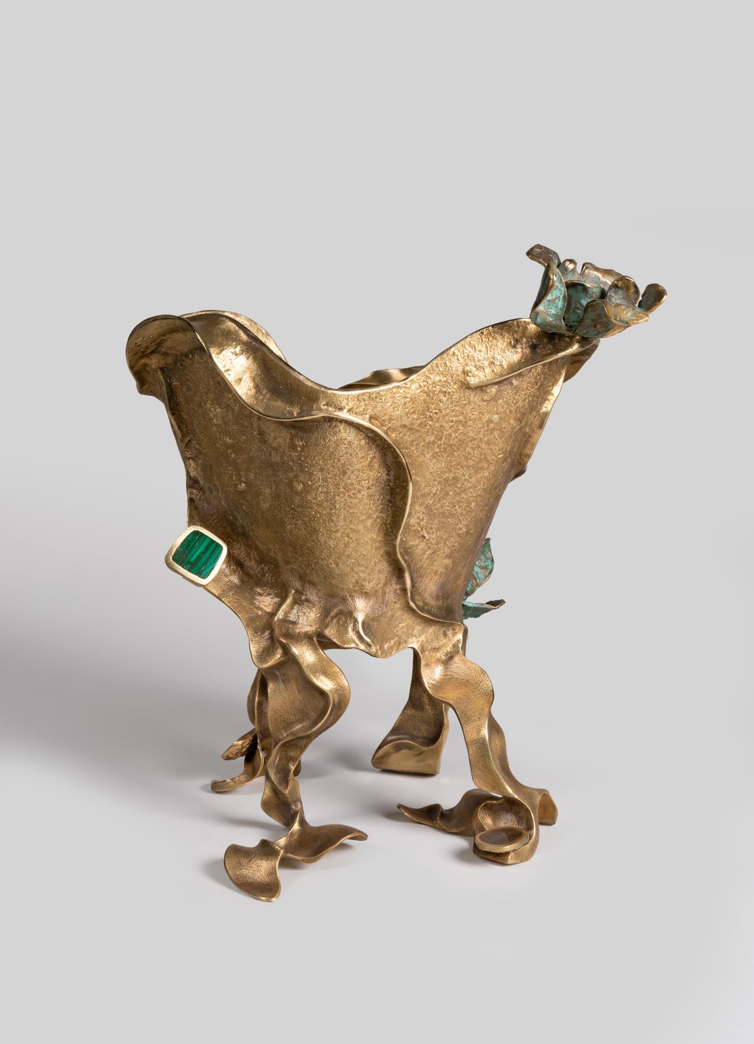 A sculptural decorative object made of textured brass with organic, flowing shapes and irregular, curling legs. It features accents of turquoise and green stones, with a prominent rectangular green gemstone set into one side.