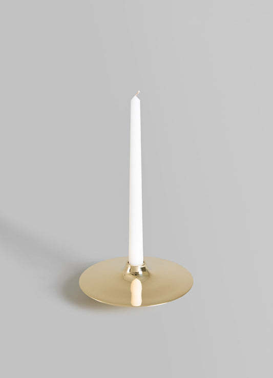 A modern candle holder with a round, flat metallic gold base and a central brass accent, holding a tall white taper candle against a neutral background.