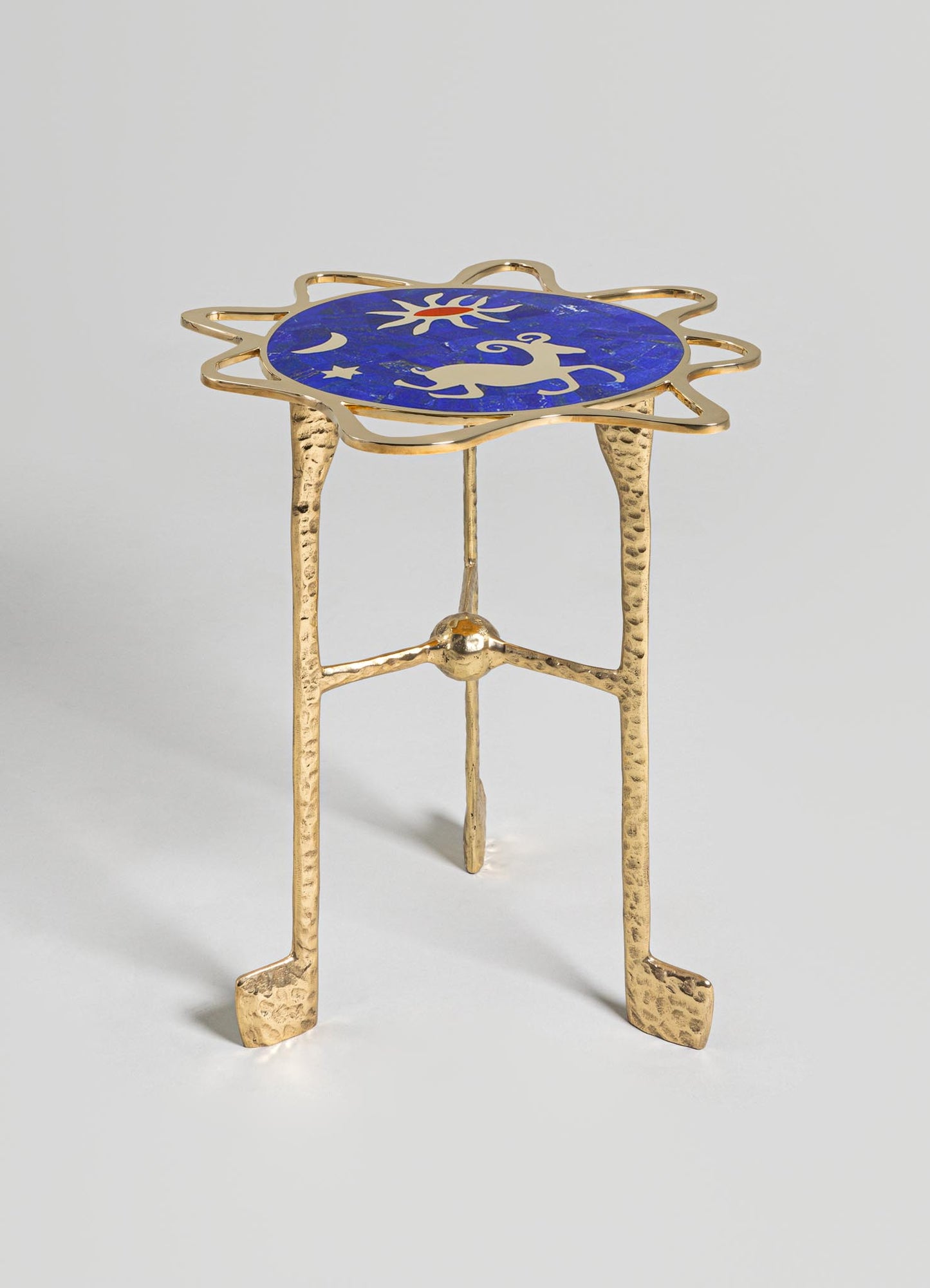 A small side table with a brass frame featuring a textured, hammered finish and three legs, topped with a circular blue mosaic surface decorated with white celestial motifs including a sun, moon, star, and reindeer.