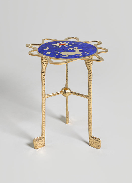 A small side table with a brass frame featuring a textured, hammered finish and three legs, topped with a circular blue mosaic surface decorated with white celestial motifs including a sun, moon, star, and reindeer.