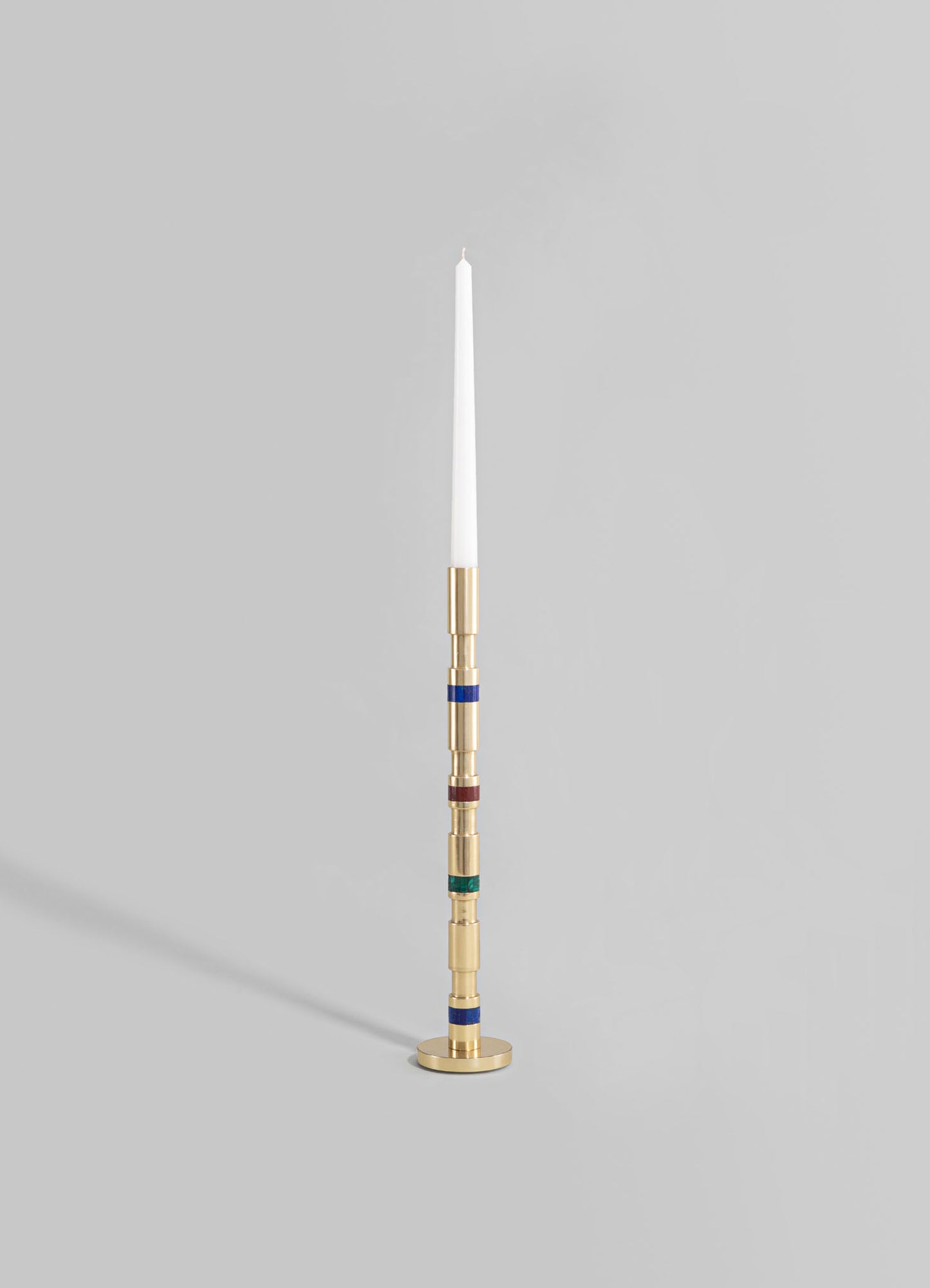 A tall candle holder with a modern design, featuring a cylindrical brass base and stem with colorful glass or enamel accents in red, blue, and green, holding a tall white taper candle.