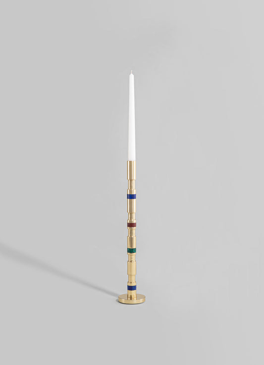 A tall candle holder with a modern design, featuring a cylindrical brass base and stem with colorful glass or enamel accents in red, blue, and green, holding a tall white taper candle.