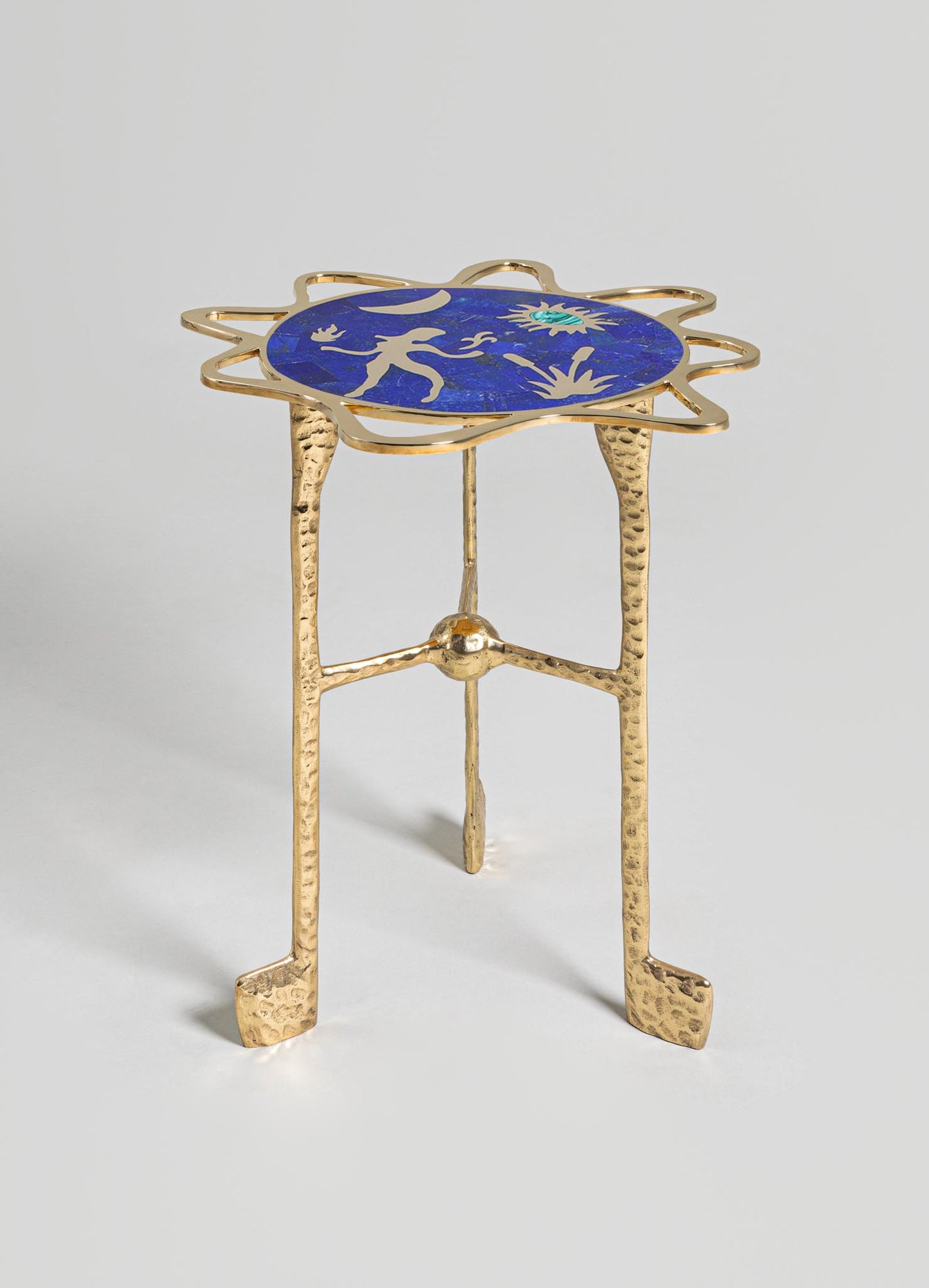 A decorative side table with a gold-toned hammered metal frame and three legs, featuring an oval top with a blue and gold abstract motif. The design combines contemporary and artisanal craftsmanship.