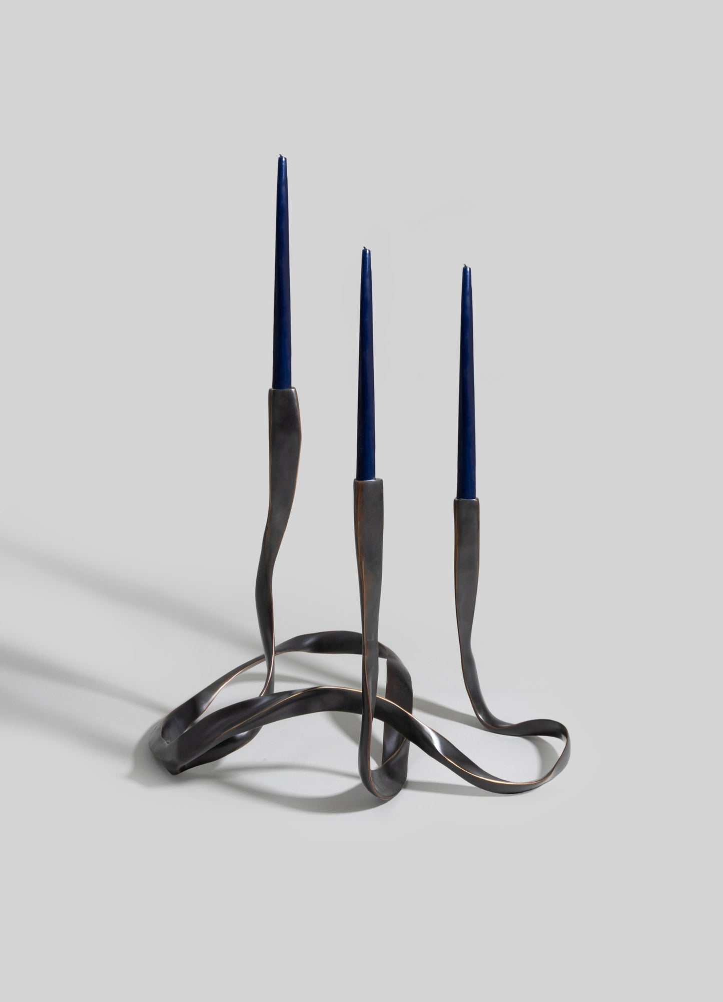 Metal candle holder with a twisted, sculptural design and a dark, matte finish, holding three tall, slender blue candles.