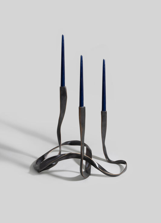 Metal candle holder with a twisted, sculptural design and a dark, matte finish, holding three tall, slender blue candles.