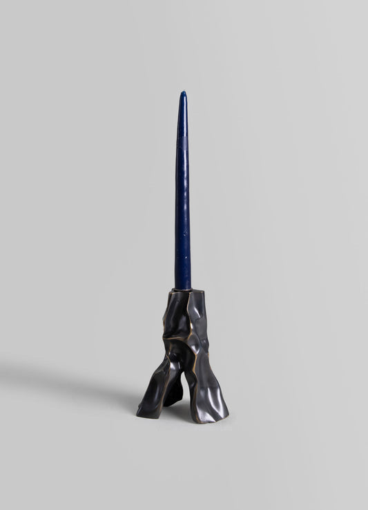 A modern candle holder with a sculptural, textured black metal base and a tall, slender dark blue candle. The design features an abstract, irregular form that emphasizes contemporary style.