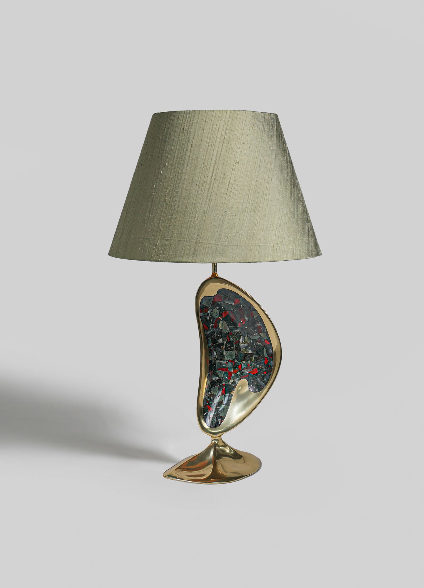 A table lamp with a ceramic base featuring an abstract, organic shape and a mosaic pattern in dark and red tones, topped with a neutral fabric lampshade.