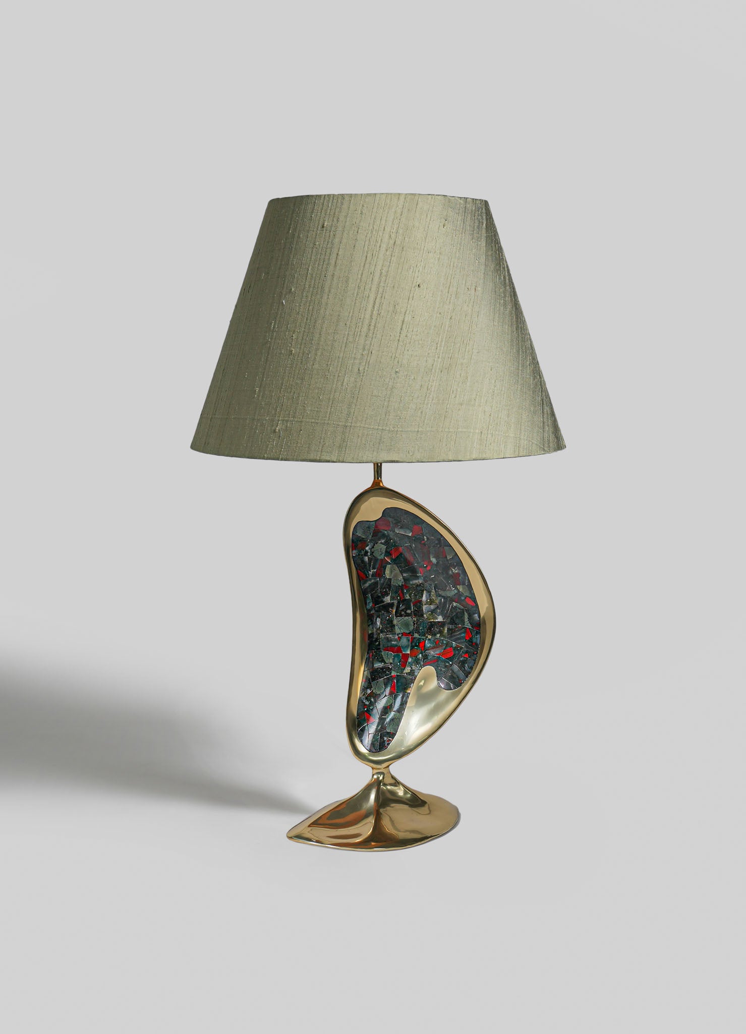 A table lamp with a ceramic base featuring an abstract, organic shape and a mosaic pattern in dark and red tones, topped with a neutral fabric lampshade.
