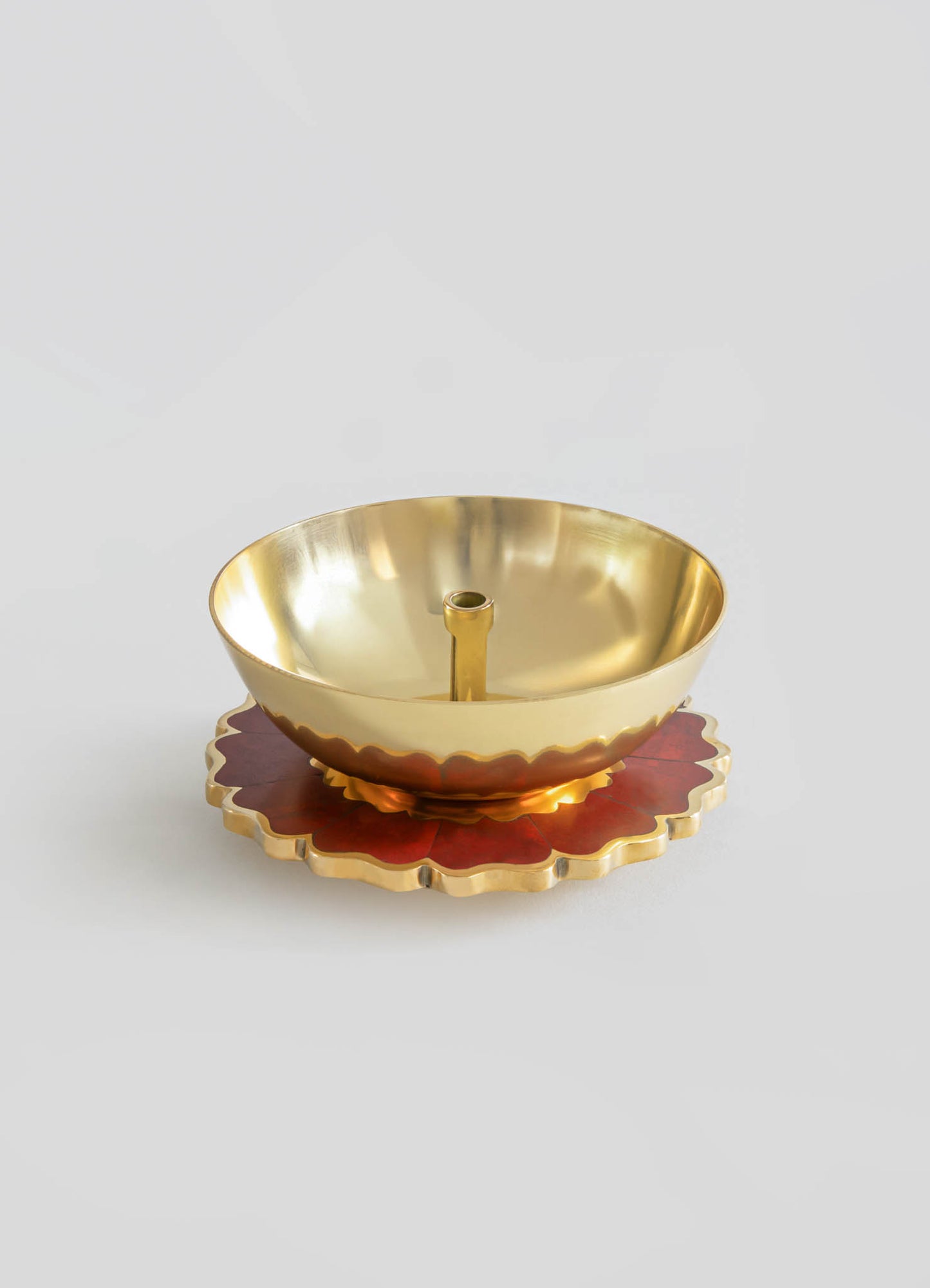 A brass candle holder with a smooth, polished surface and a central socket for a candle, placed on a decorative red and gold scalloped tray with a floral motif.