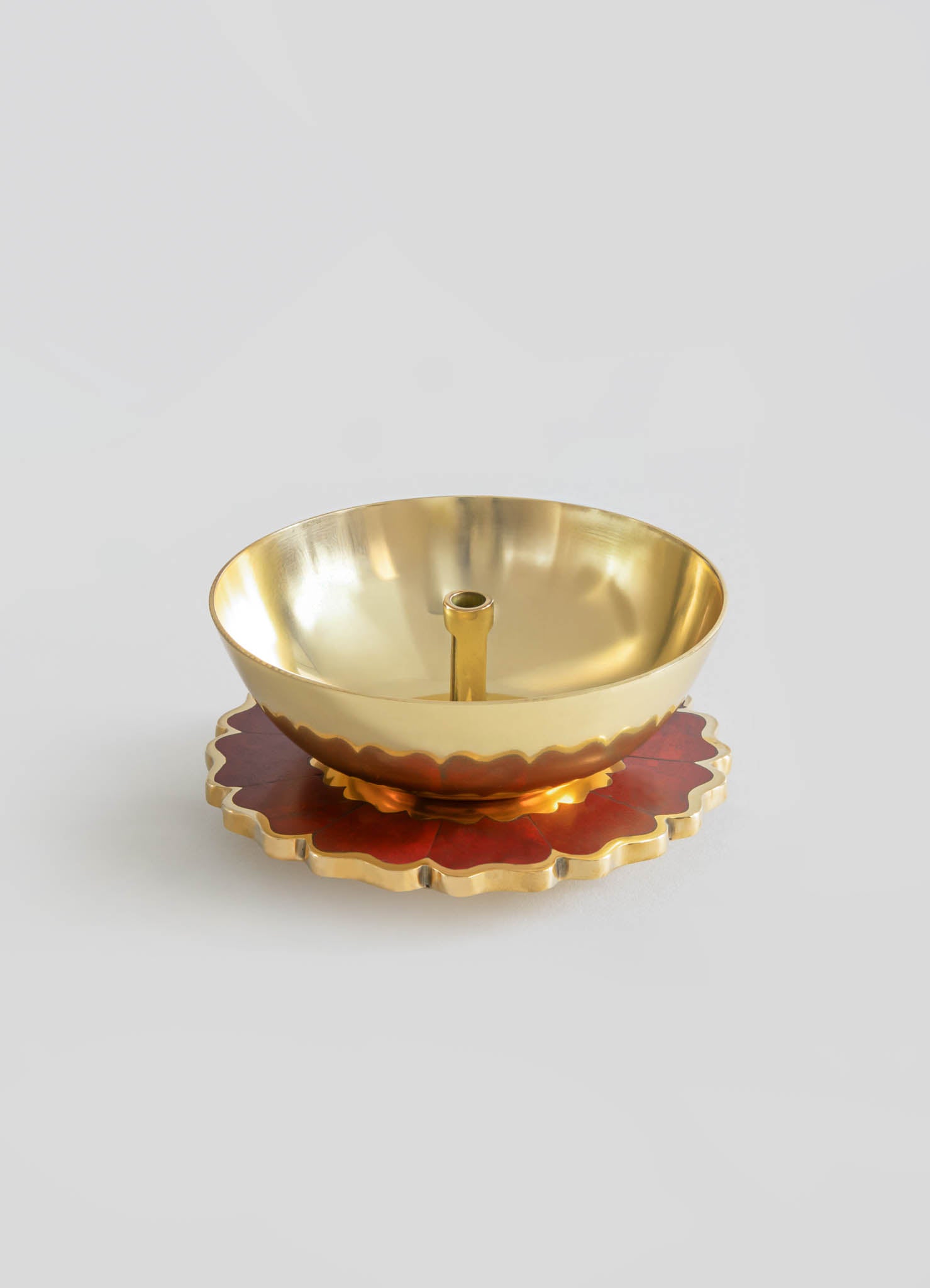 A brass candle holder with a smooth, polished surface and a central socket for a candle, placed on a decorative red and gold scalloped tray with a floral motif.