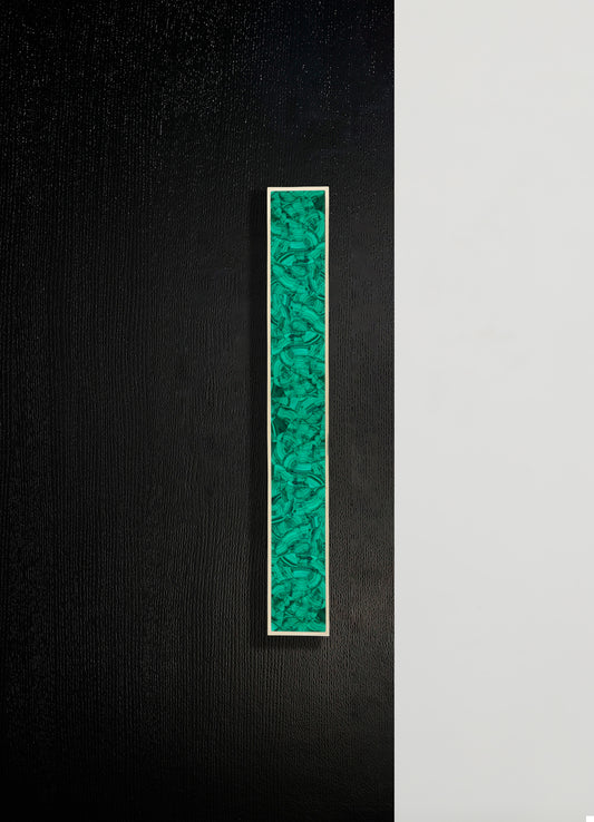 A long rectangular artwork with an abstract pattern in shades of green and black, framed with a thin white border, mounted on a textured black wall.