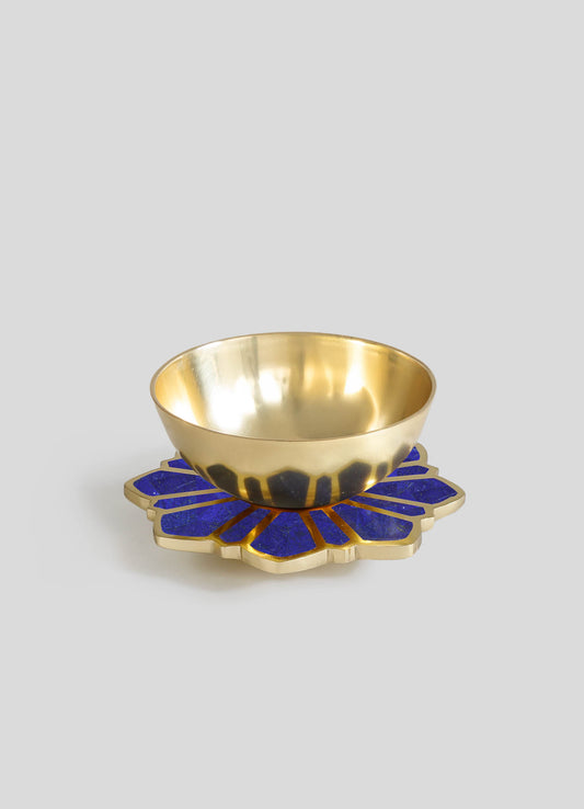 A polished brass bowl with a smooth, reflective surface rests on a decorative base featuring deep blue inlay with a floral or petal pattern, combining metallic and colored materials.