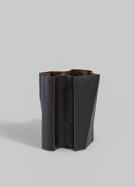 A black metal vase with a matte, textured finish featuring an irregular, geometric shape with vertical ridges. The design is contemporary and minimalist, showcasing a sturdy, sculptural form.