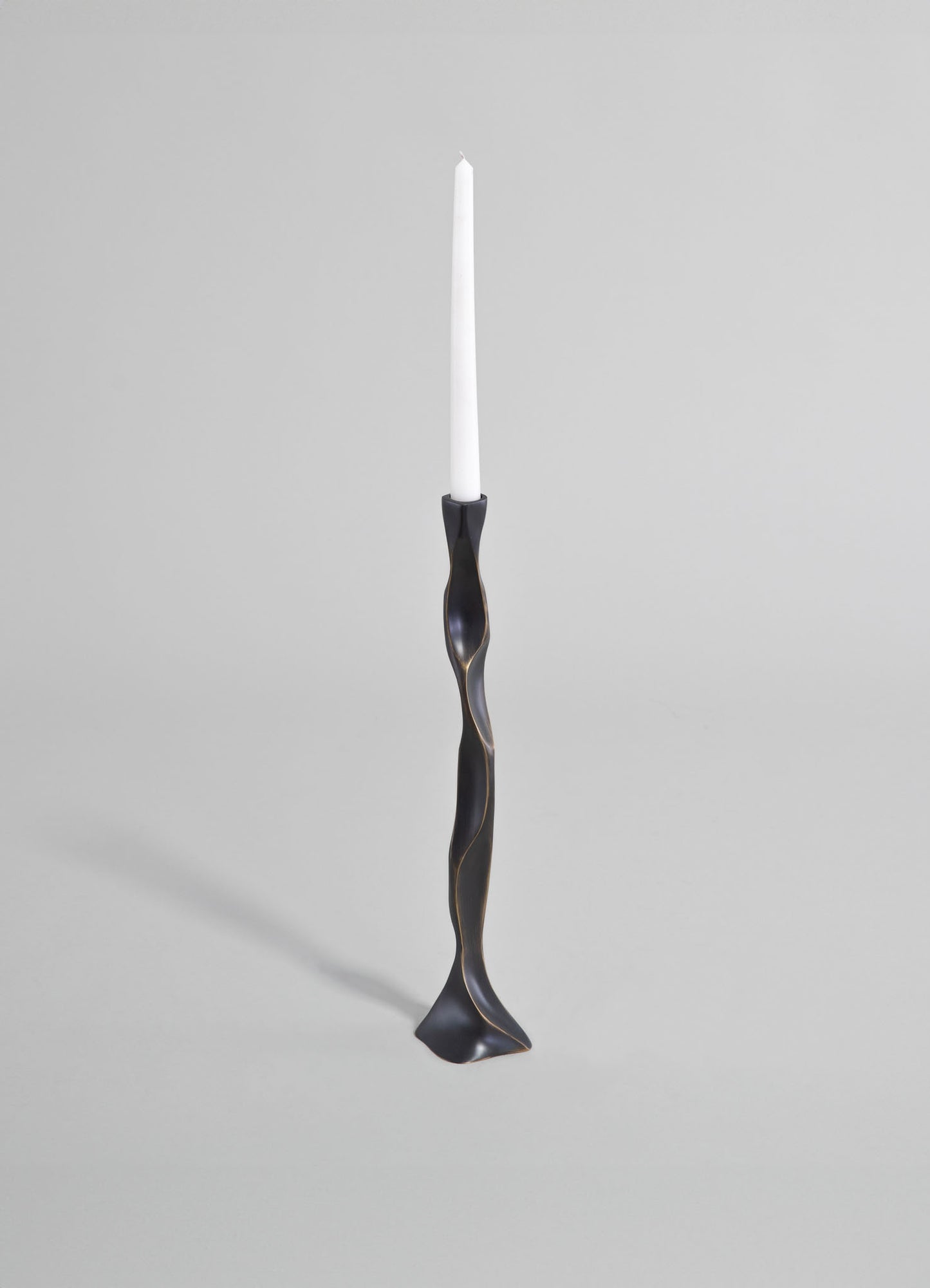 A sculptural candlestick holder made of black metal with a twisted, organic shape and a smooth matte finish, holding a white taper candle against a plain gray background.