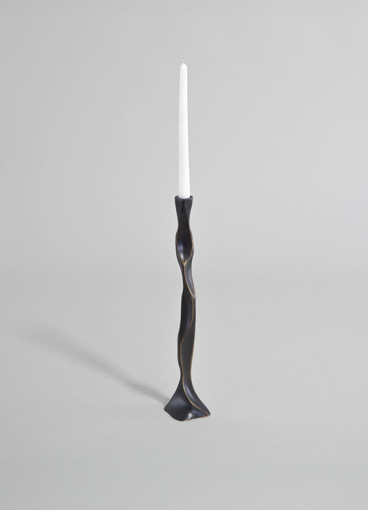A sculptural candlestick holder made of black metal with a twisted, organic shape and a smooth matte finish, holding a white taper candle against a plain gray background.