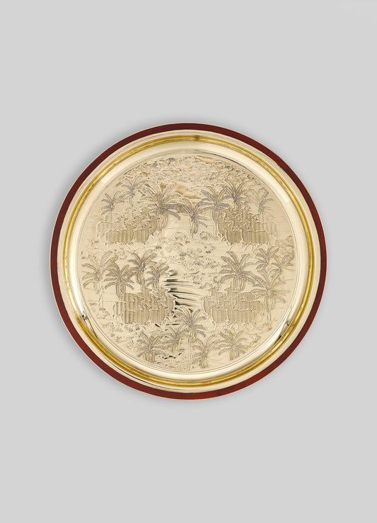 A decorative round tray with a polished gold finish featuring an engraved tropical landscape motif with palm trees and foliage, bordered by a dark wood rim.