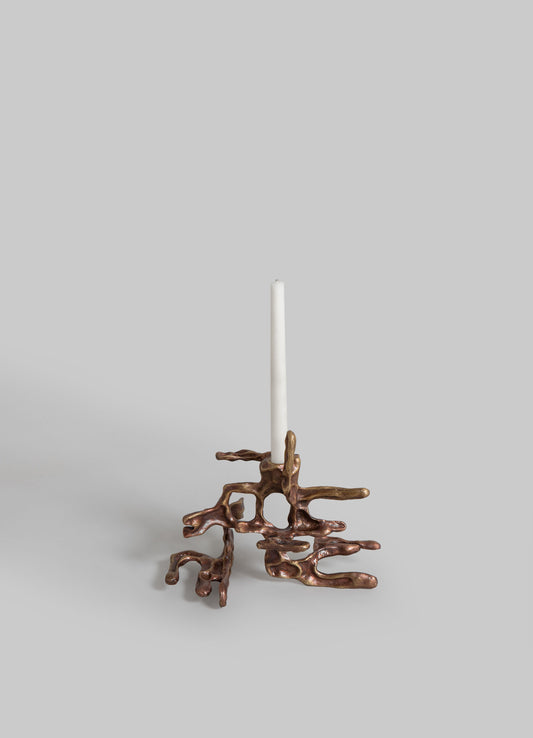 A candle holder with a sculptural, organic brass design resembling intertwined roots or branches, supporting a tall white candle against a plain gray background.