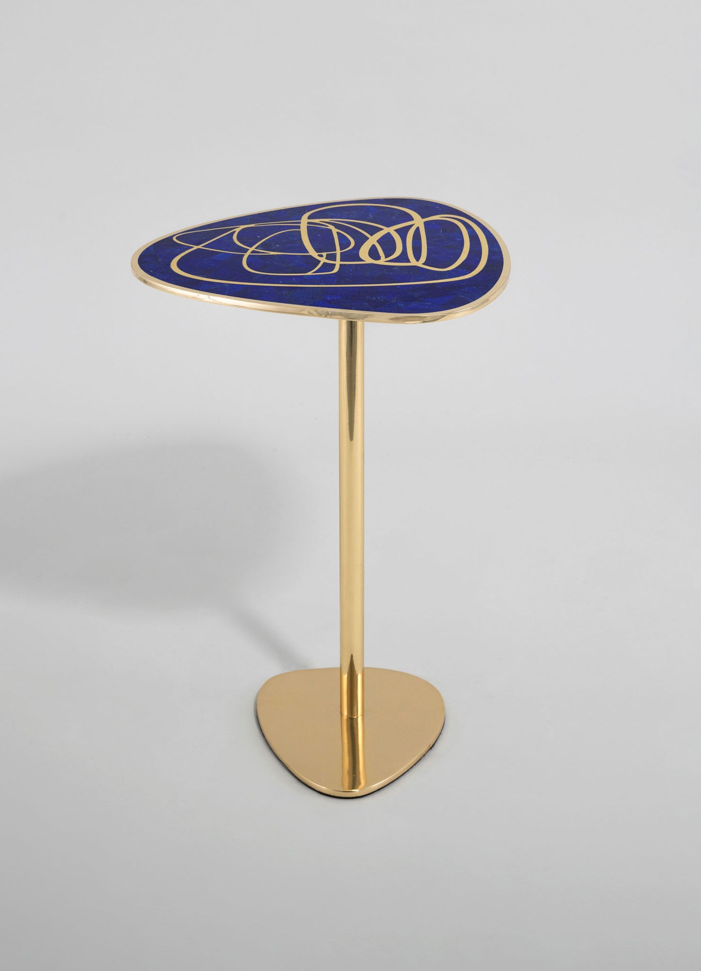 A modern side table with a triangular-shaped top featuring a blue and gold abstract pattern, supported by a gold metallic stand with an oval base. The design combines contemporary style with luxurious accents.