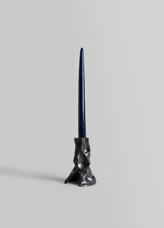 A tall, sculptural candlestick holder made of black ceramic with organic, irregular contours and a matte finish, holding a slender, tapered dark blue candle.