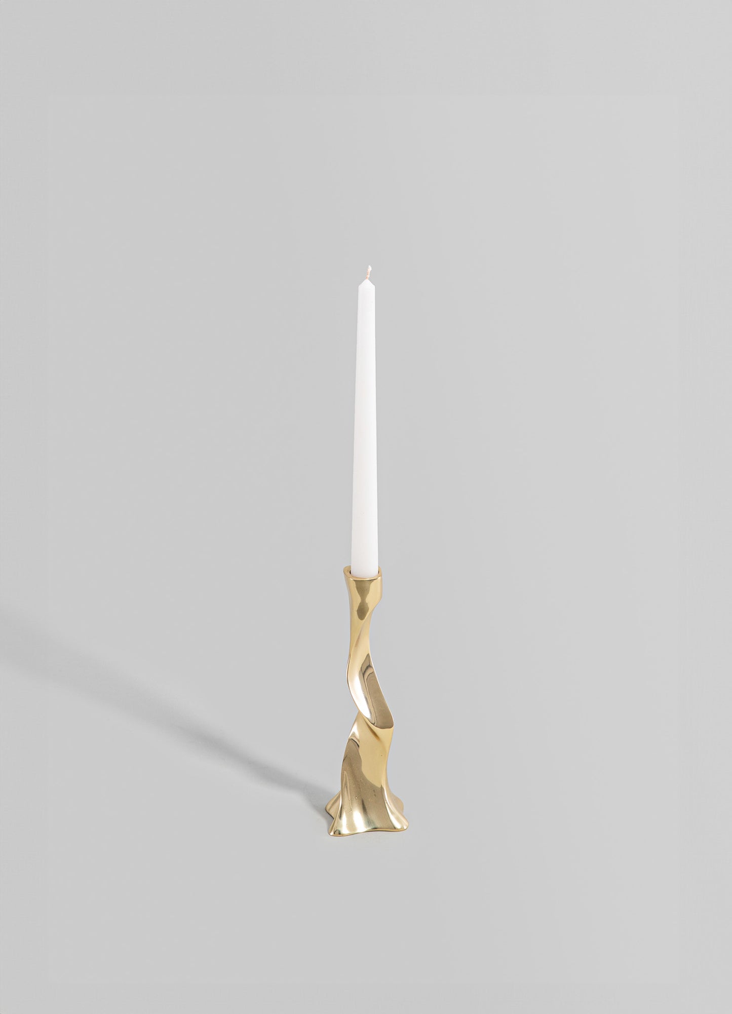A brass candle holder with an abstract, sculptural form and a polished metallic finish, holding a tall white taper candle. The design is contemporary with organic curves and a minimalist aesthetic.