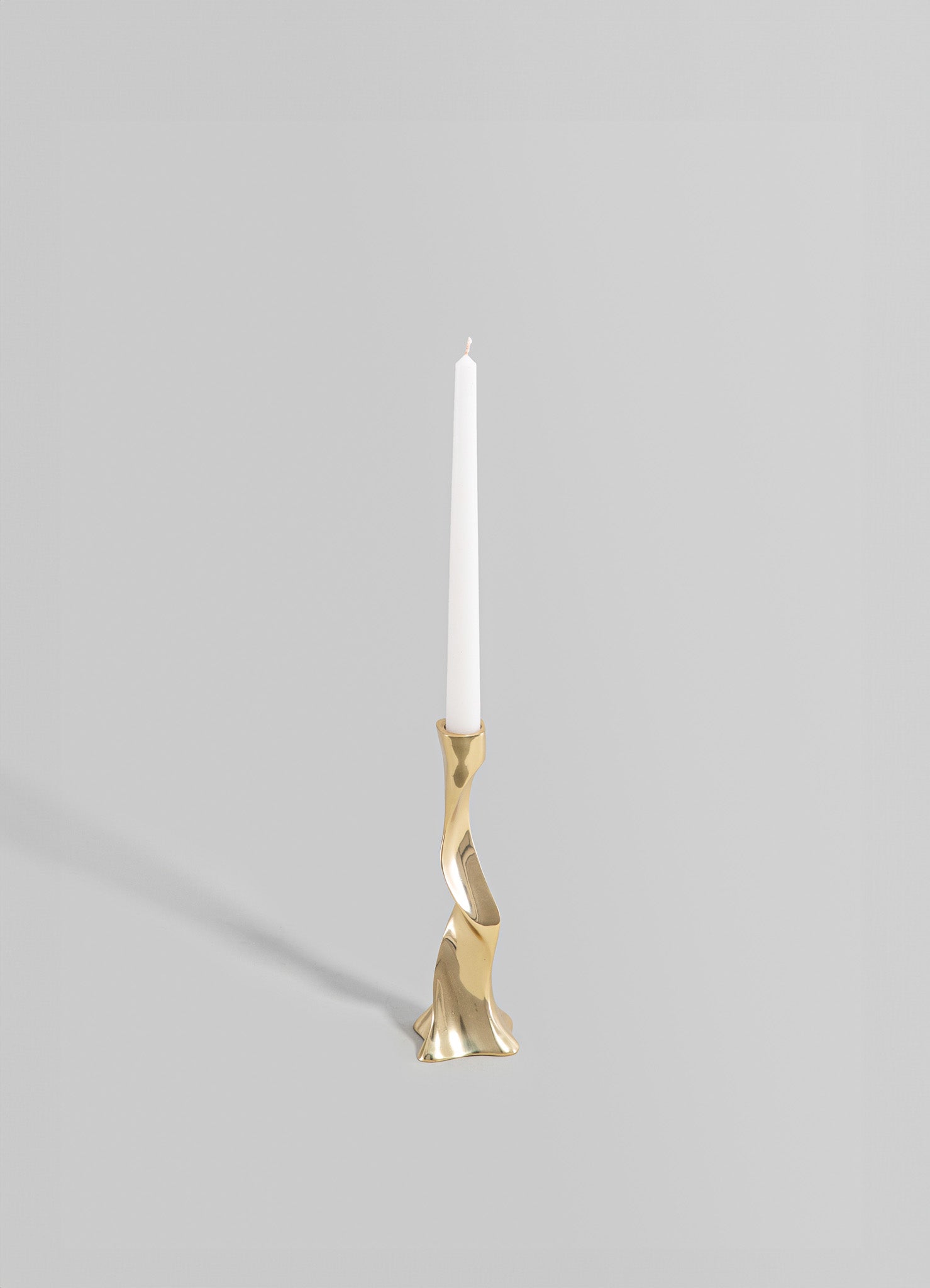 A brass candle holder with an abstract, sculptural form and a polished metallic finish, holding a tall white taper candle. The design is contemporary with organic curves and a minimalist aesthetic.