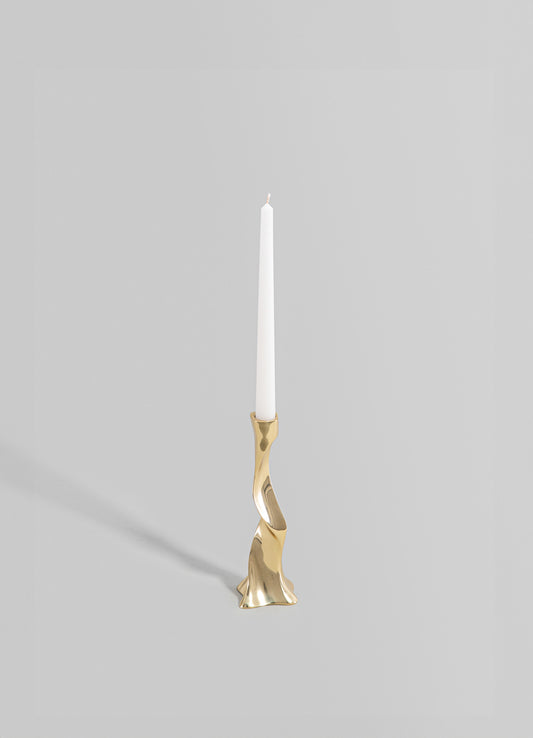 A brass candle holder with an abstract, sculptural form and a polished metallic finish, holding a tall white taper candle. The design is contemporary with organic curves and a minimalist aesthetic.
