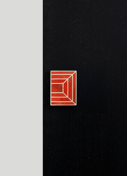 A rectangular pin with a geometric pattern in red and white, placed on a black textured surface with subtle dotted lines.