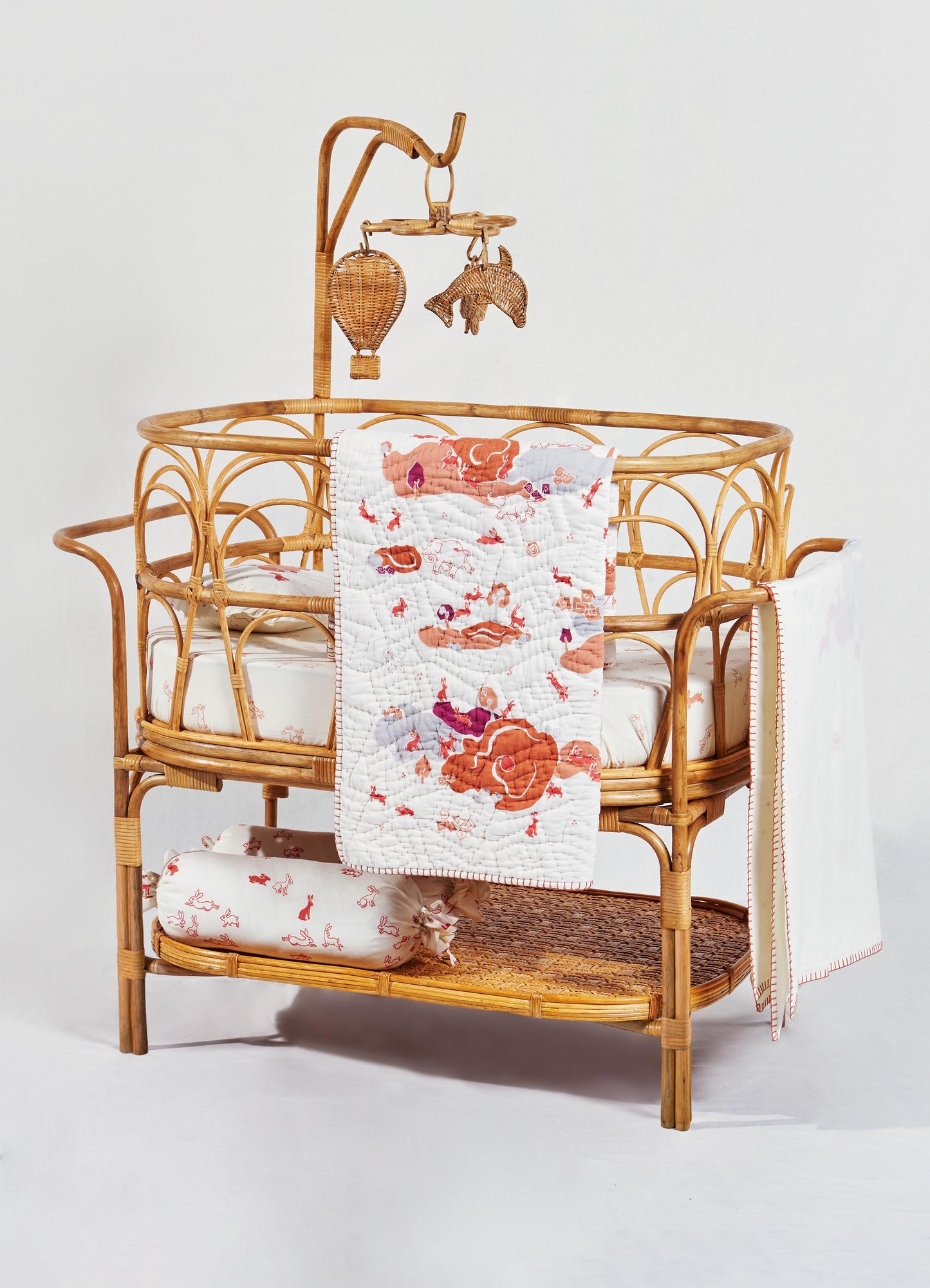 A rattan crib with a natural finish, featuring intricate curved and woven details, decorated with white fabric bedding and a quilt with floral and animal motifs in shades of orange and purple. A mobile with wicker hot air balloon, horse, and animal shapes hangs above the crib.