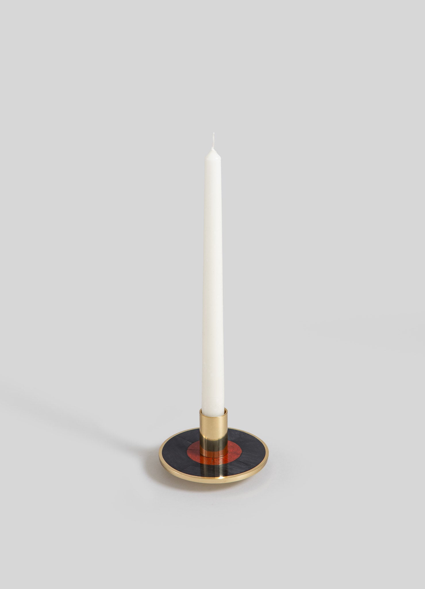 A tall white candle on a decorative candlestick holder with a round base featuring black, red, and gold accents, and a metallic gold band around the candle. The design combines contemporary with minimalist style.