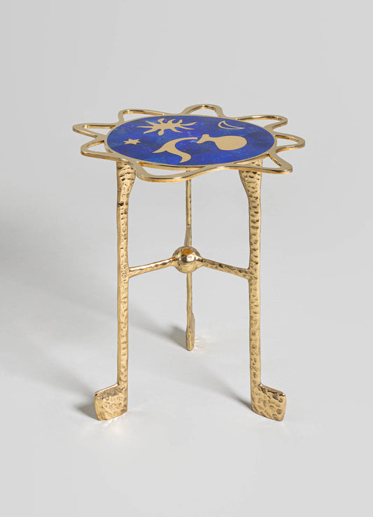 A small side table with a gold hammered metal frame and three slender legs, topped with an oval-shaped inlay featuring a vibrant blue and gold abstract artistic motif.