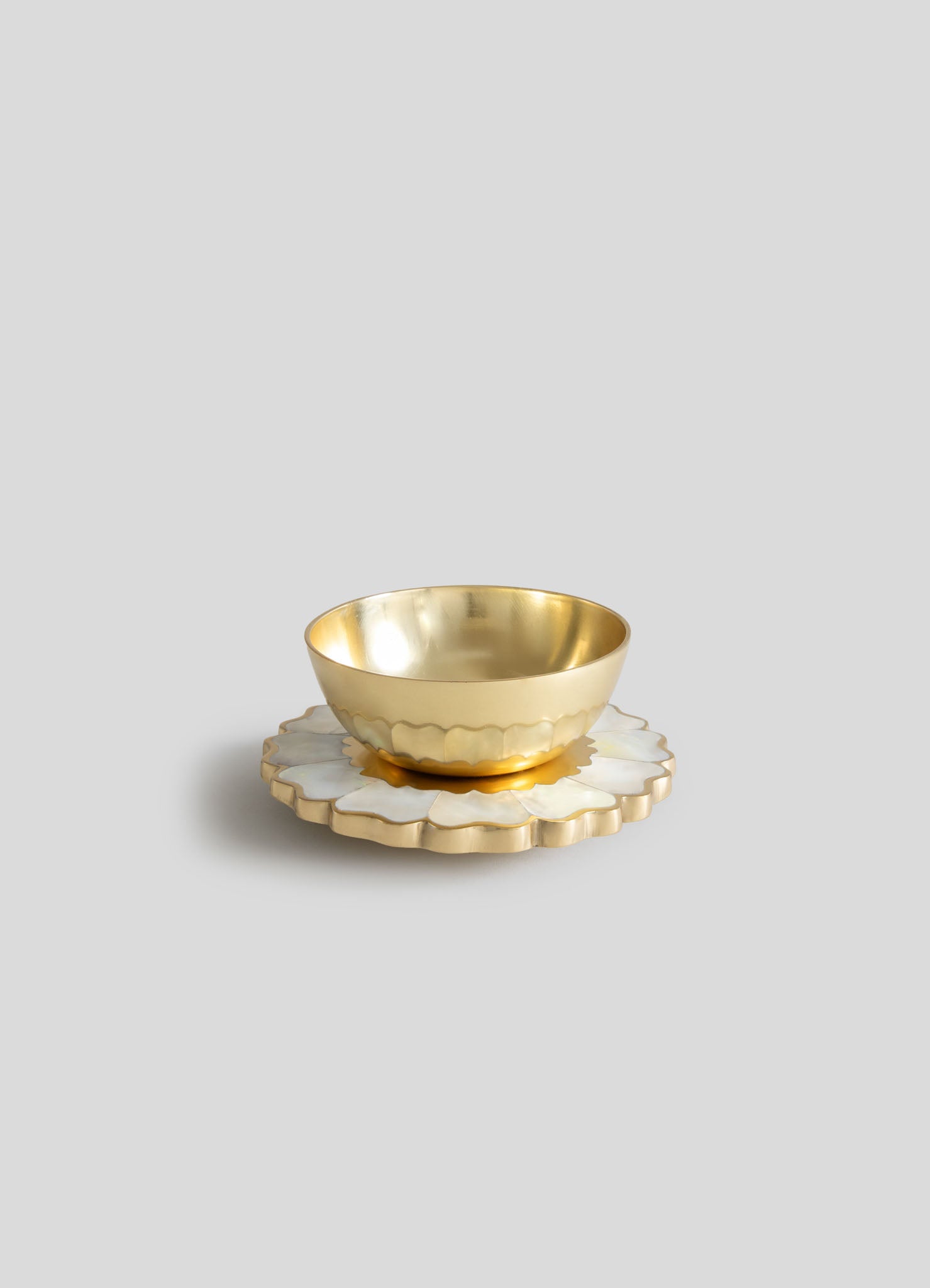 A gold-colored metal bowl with a polished finish, placed on a white and gold scalloped ceramic coaster with a floral motif.