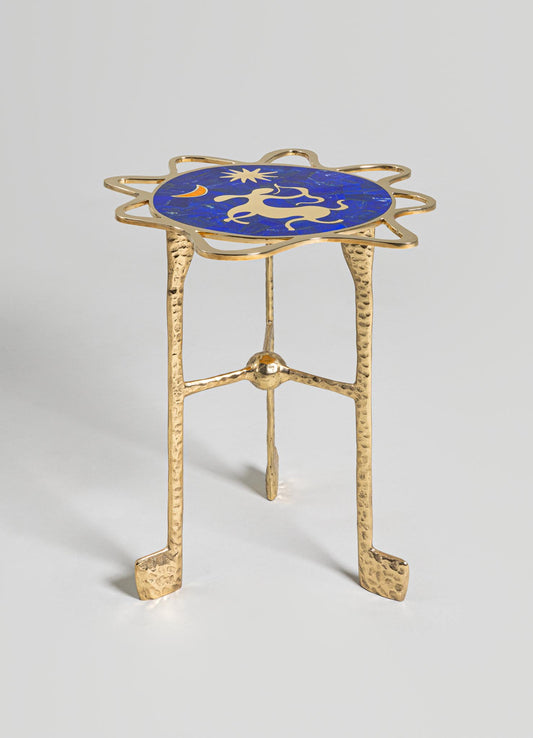 A small side table with a gilded brass base featuring textured, hammered legs and a circular top inlaid with blue lapis lazuli, decorated with celestial motifs including a sun, moon, and star patterns.