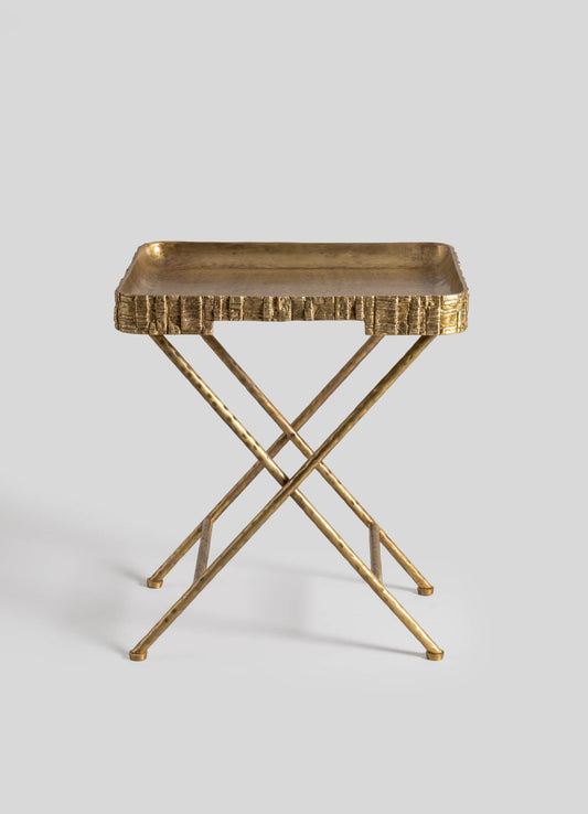 A small accent table with a rectangular brass tray top featuring a textured edge design, supported by crossed, slender brass legs with a polished finish.
