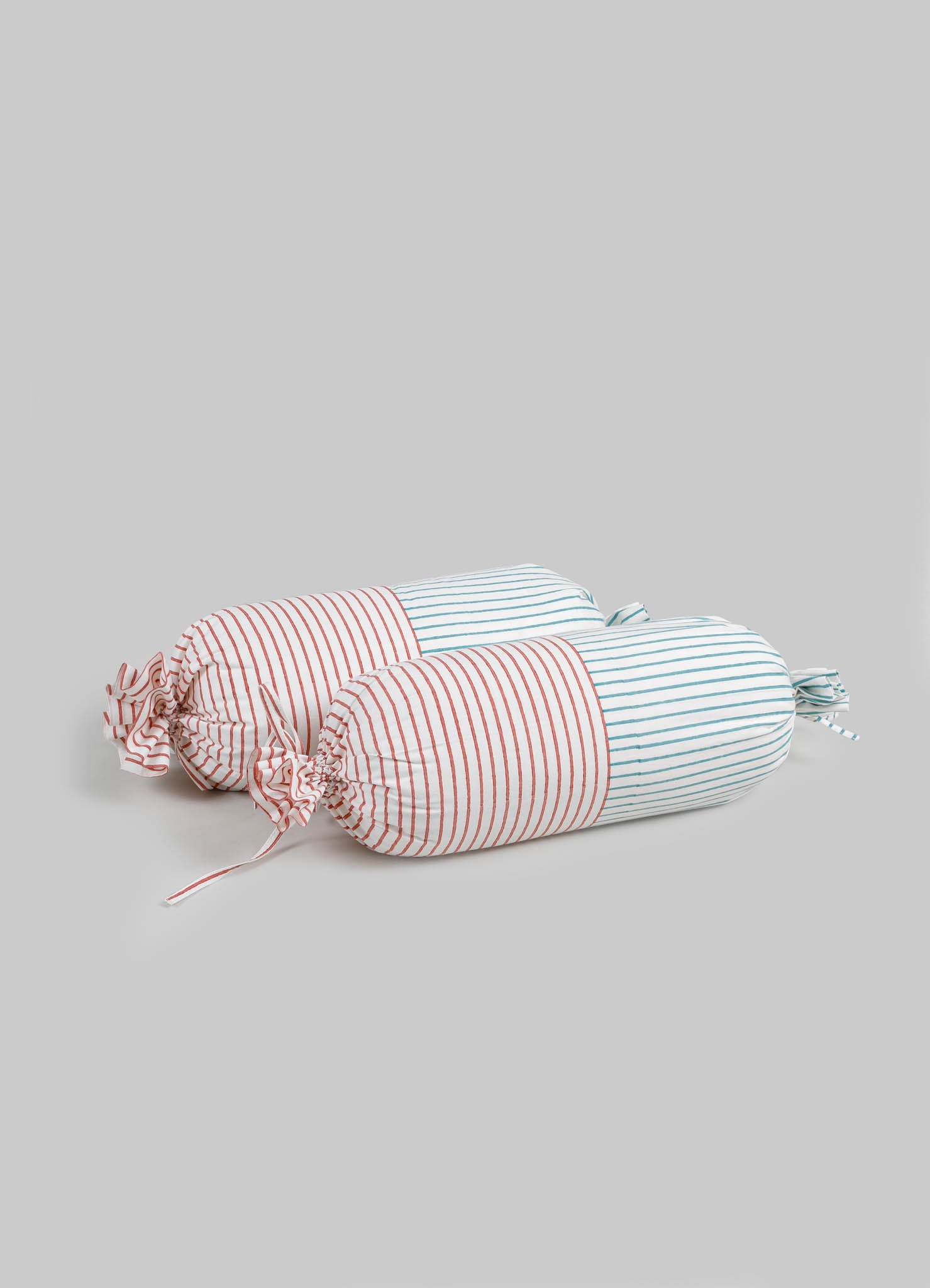 Striped fabric bolsters with red and blue thin lines on white background, featuring tied fabric ends. The cushions have a soft, padded appearance and a casual, handcrafted style.