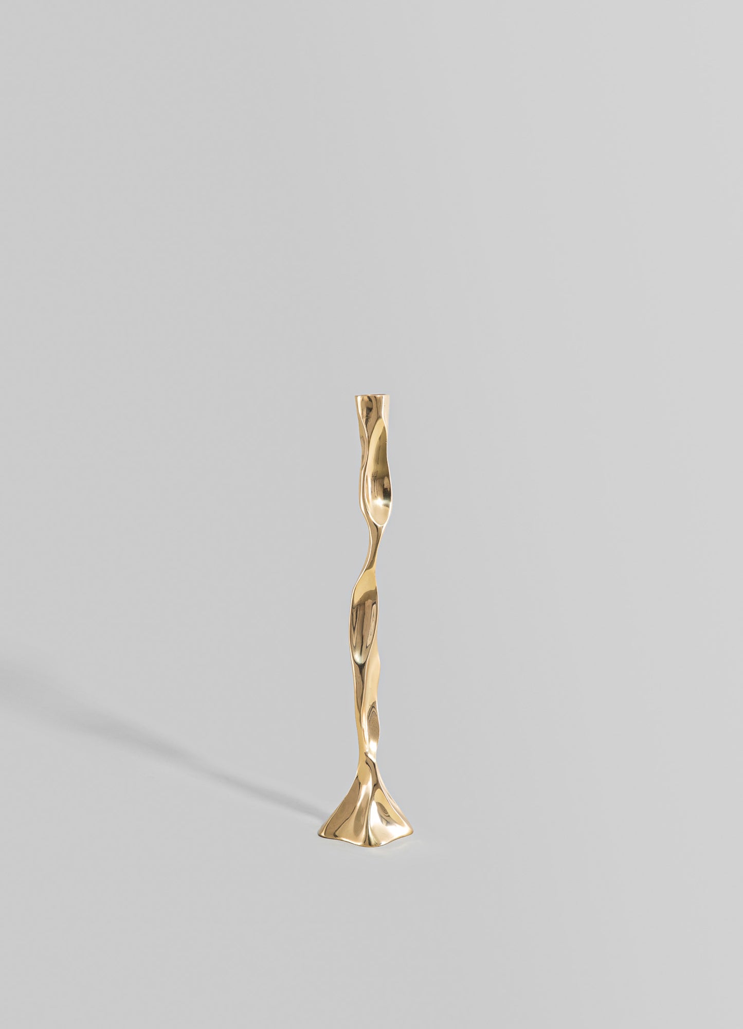 A tall, sculptural brass candle holder with an irregular, twisted form and a polished gold finish, set against a light gray background.