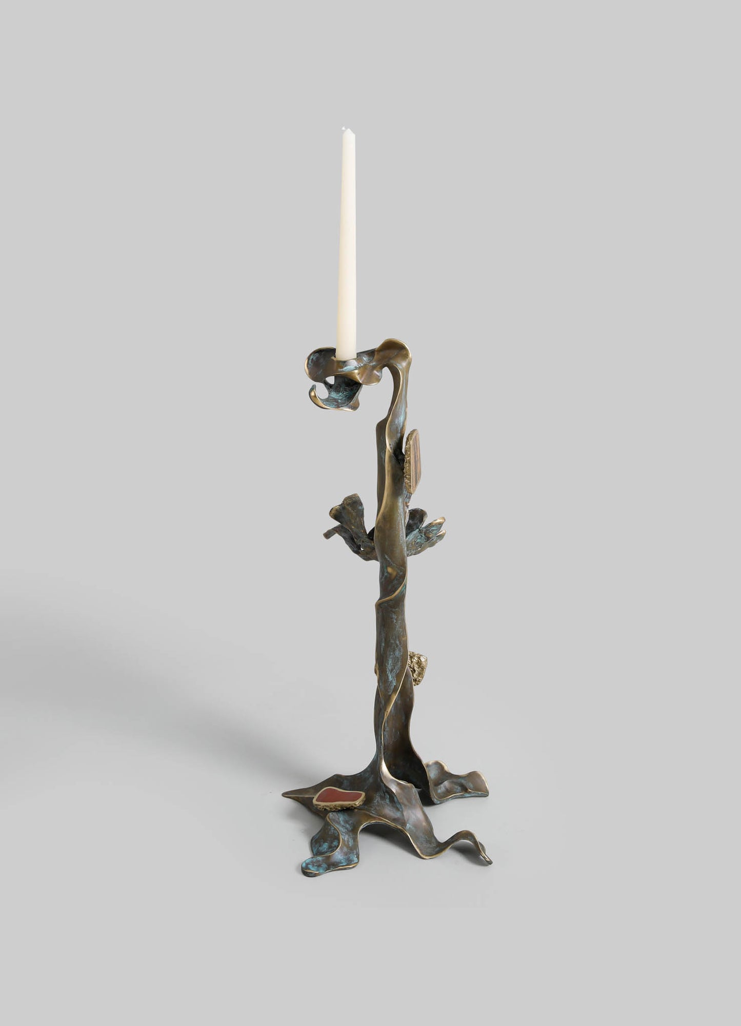 A brass candlestick with an organic, abstract design resembling twisted natural forms, featuring a tall, white taper candle. The surface has a textured, antique finish with subtle patinas and irregular edges.