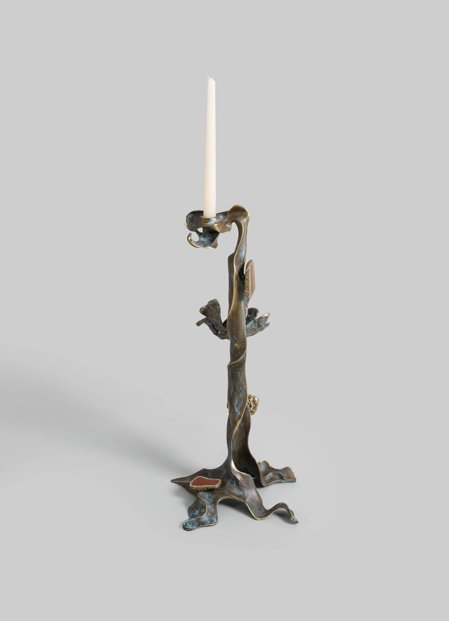 A brass candlestick with an organic, abstract design resembling twisted natural forms, featuring a tall, white taper candle. The surface has a textured, antique finish with subtle patinas and irregular edges.