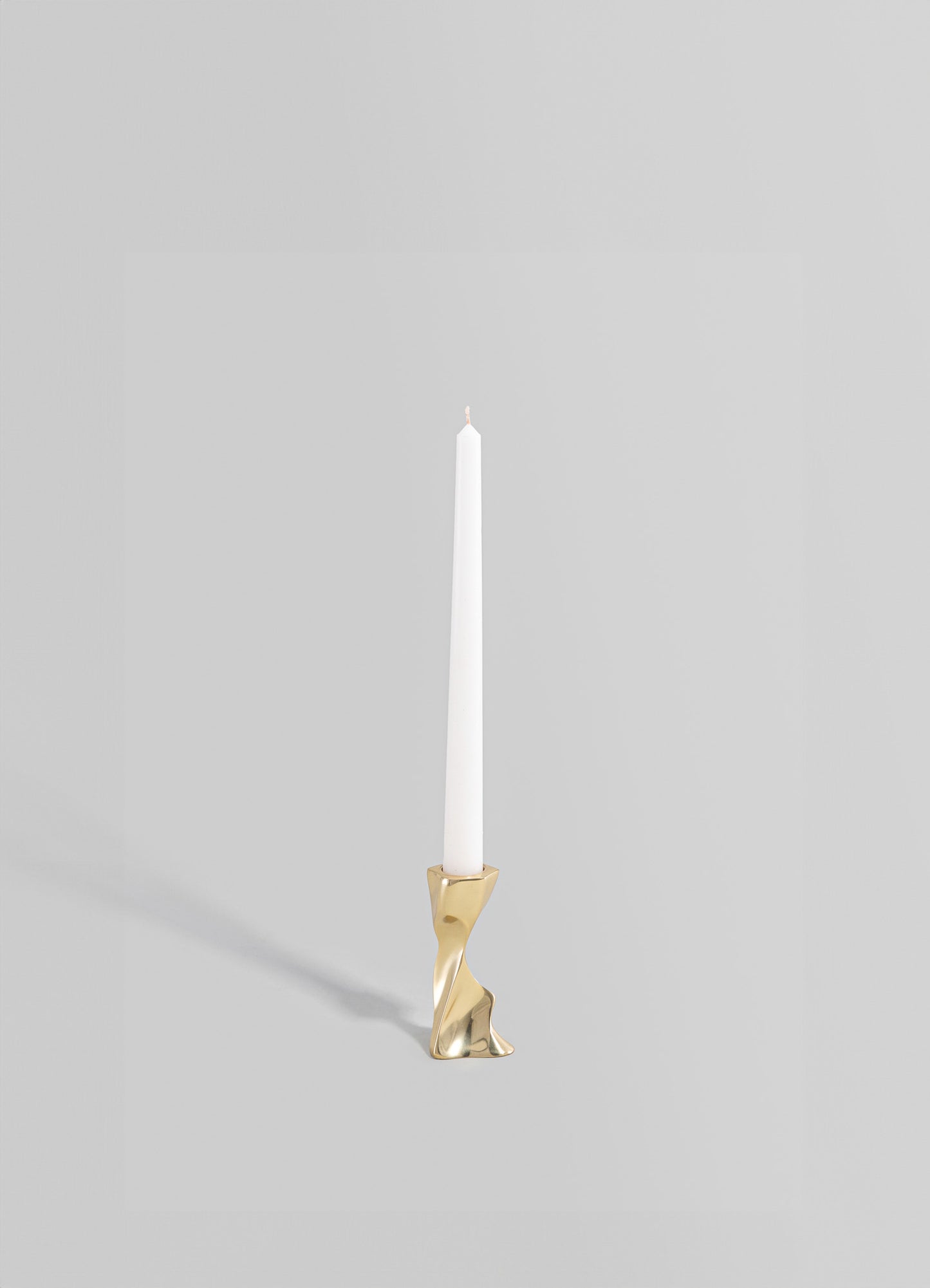 A brass candle holder with an irregular, sculptural shape, holding a white taper candle. The finish is polished and reflective, conveying a contemporary and minimalist style.
