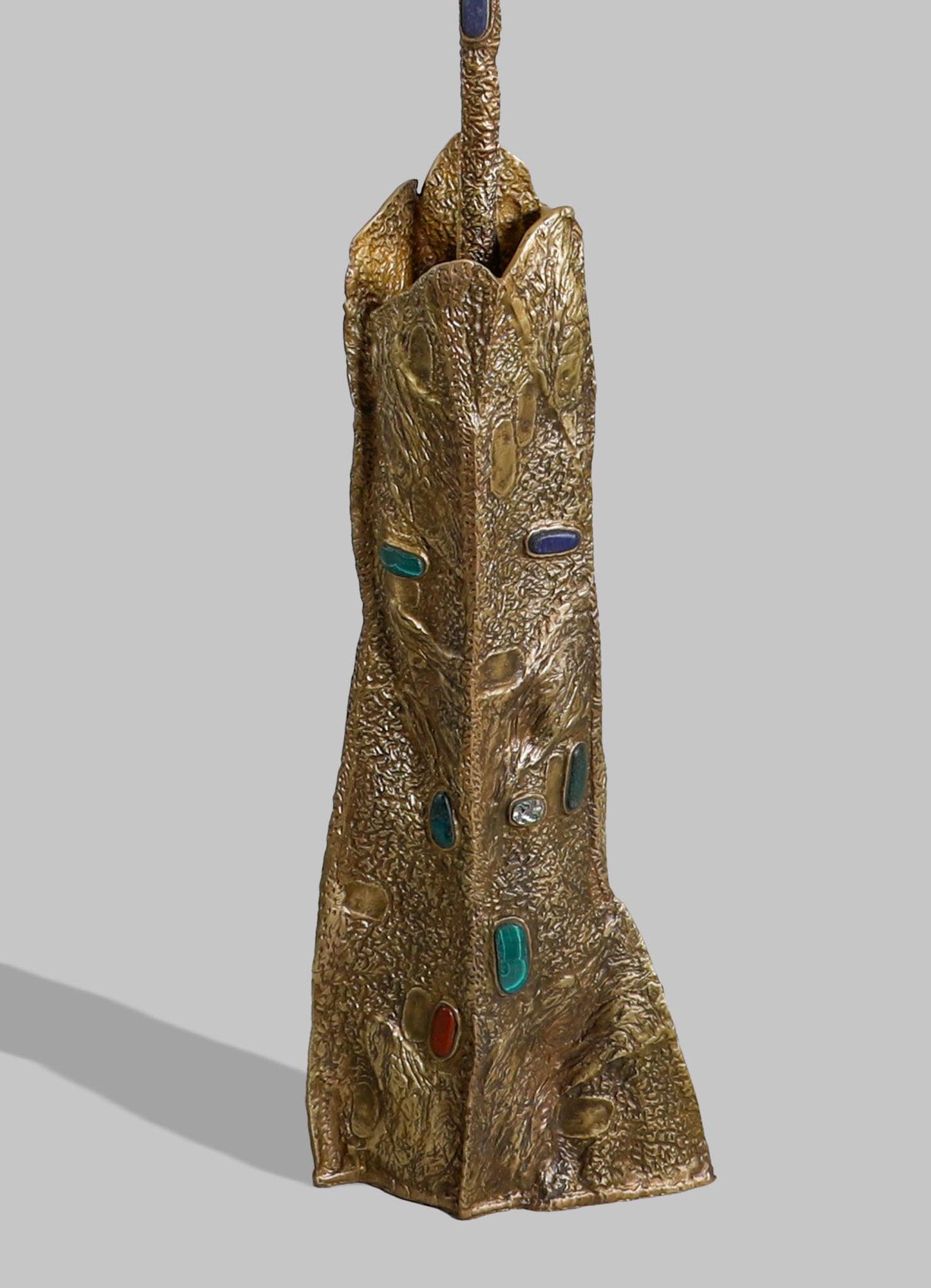 A tall decorative sculpture with a textured gold-finished surface shaped like an elongated animal head, adorned with colorful gemstone inlays in blue, green, and clear tones. The piece has a handcrafted, artistic style with a rustic and luxurious aesthetic.