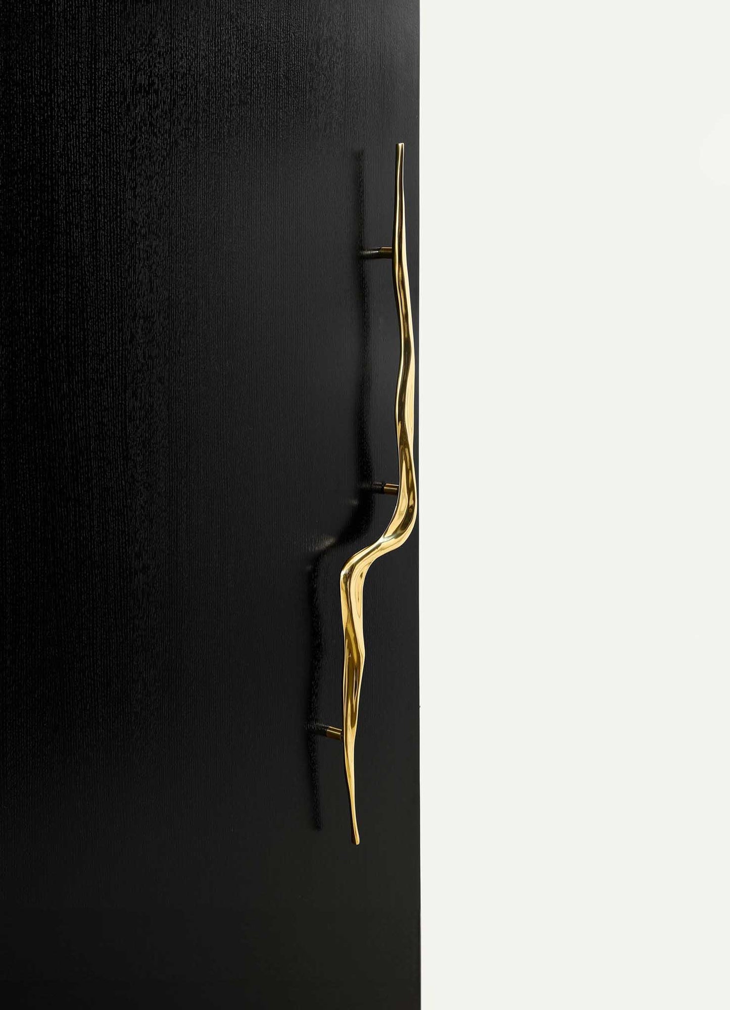 A modern gold-toned wall hook with an abstract, wavy design mounted on a black-textured wall. The hook has a sleek, contemporary style with a polished finish.