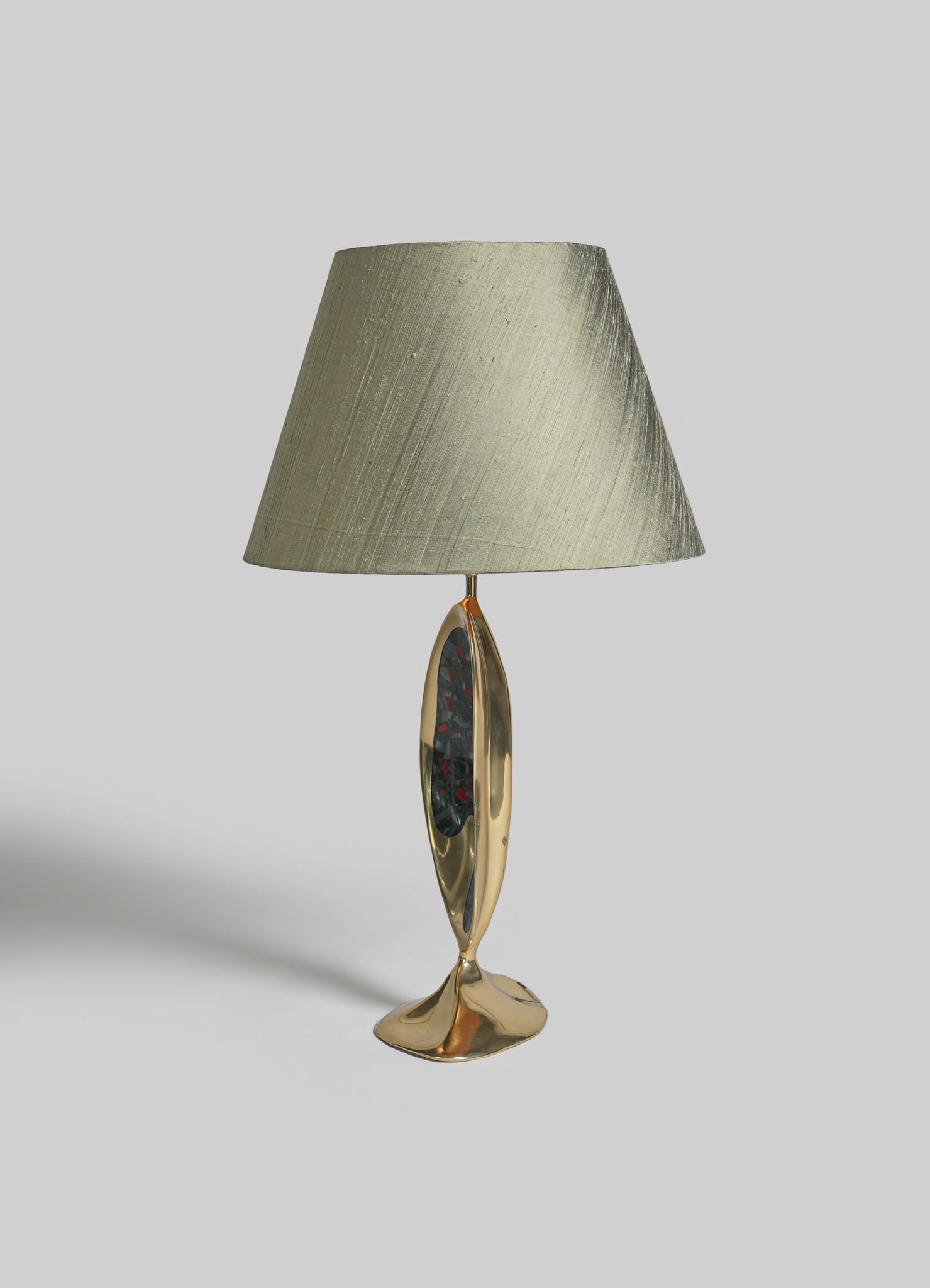 A table lamp with a beige fabric lampshade and a sculptural gold metal base featuring an oval, multicolored resin inlay. The design combines contemporary elegance with artistic detailing.