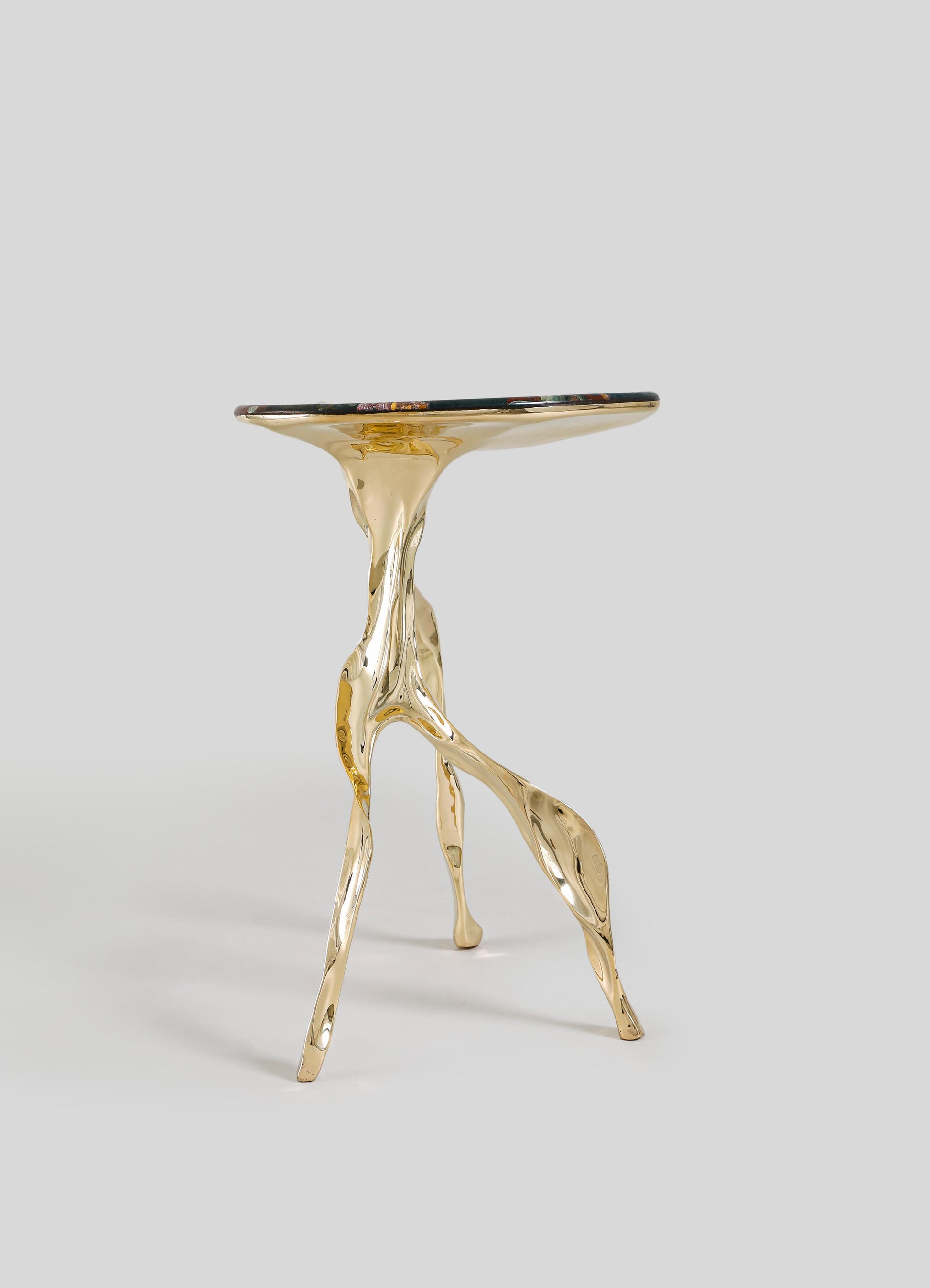 The VIYA Under The Banyan Gold Side Table Large features a sculptural gold base with three legs inspired by aerial roots, topped with a round dark glass surface, shown against a plain light gray background.