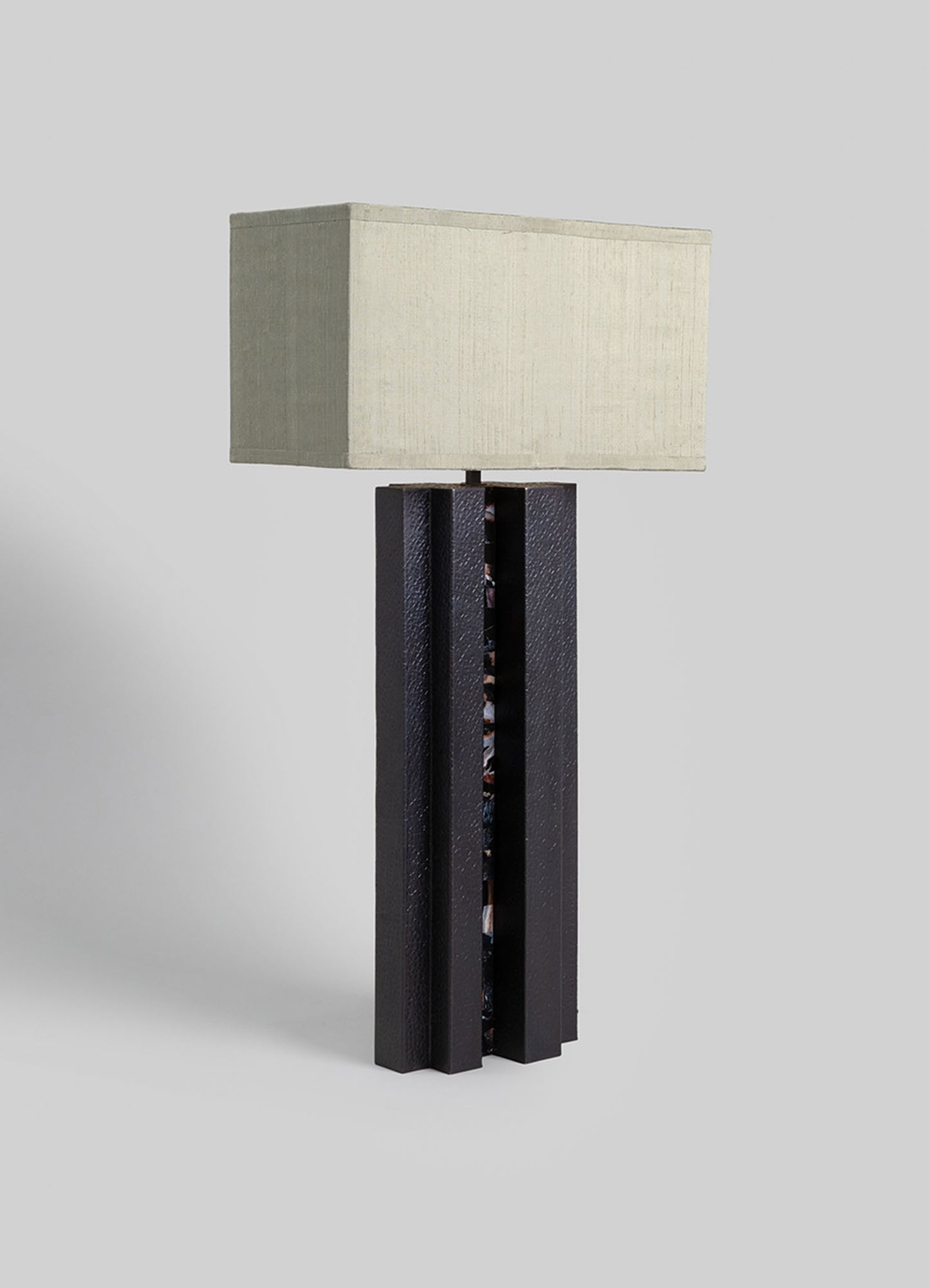 A table lamp with a rectangular fabric shade in a neutral beige tone and a textured black ceramic base featuring vertical ridges. The lamp has a contemporary design with a combination of smooth and textured materials.