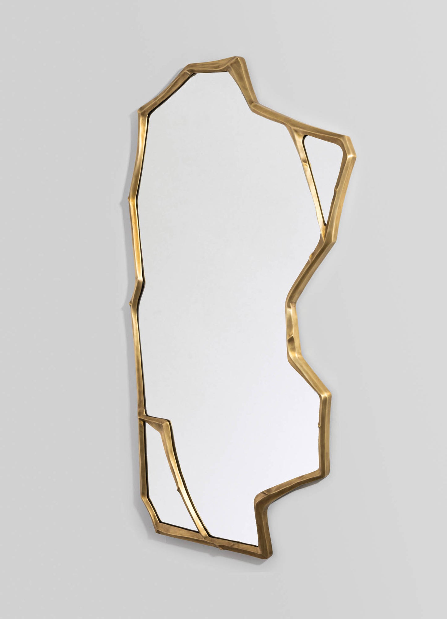 A decorative wall mirror with a frame made of irregularly shaped, metallic gold metal, creating an abstract, contemporary design. The mirror has a slim profile and is mounted on a neutral, light-colored wall.