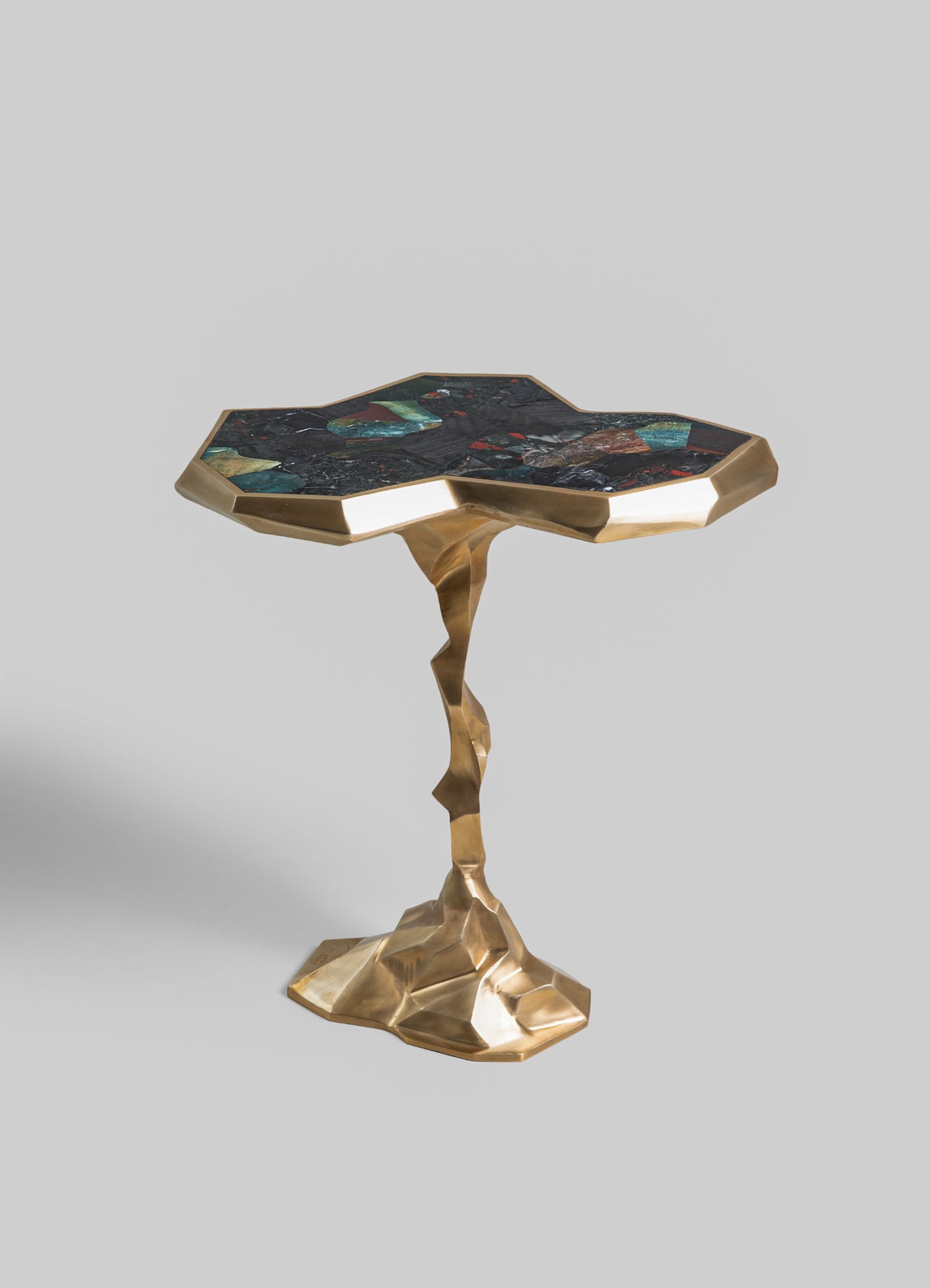 A small side table with a sculptural, irregular metal base in a gold finish and a geometric, multicolored stone top featuring dark green, black, and red tones.